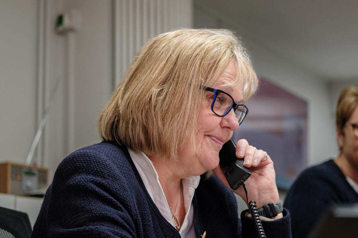 A candid shot of Tina, a receptionist at Cricklade Dental Practice, wearing glasses and a dark blue jacket, speaking on the telephone at the reception desk.