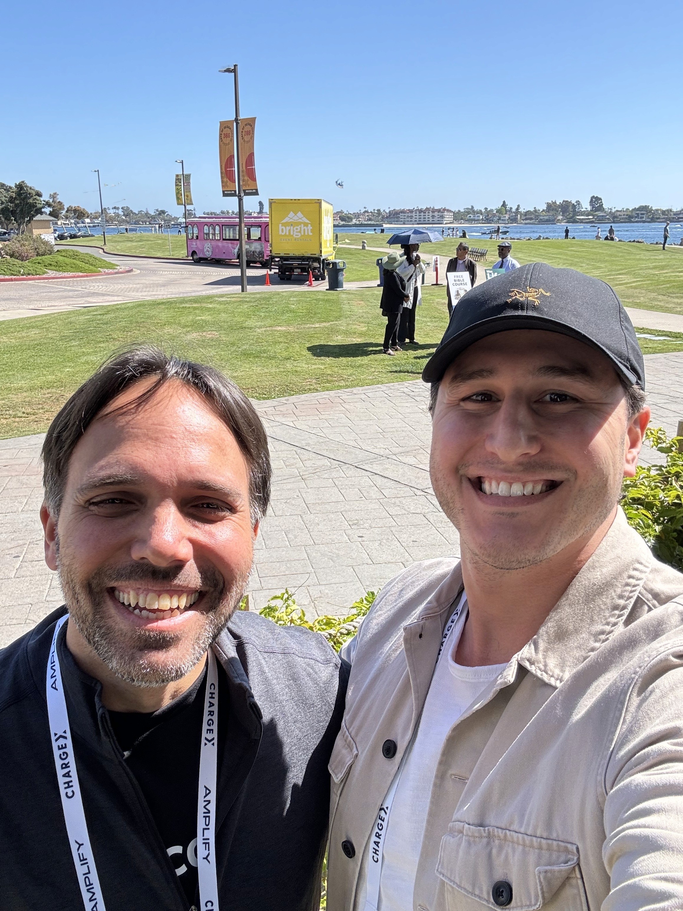 Two smiling individuals posing for a selfie outdoors, with a park and a red structure in the background.