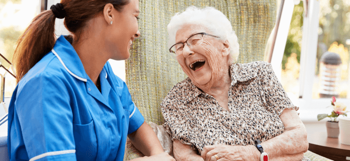 A nurse in a blue uniform shares a warm, joyful laugh with an elderly woman with white hair and glasses, in what appears to be a care home setting.