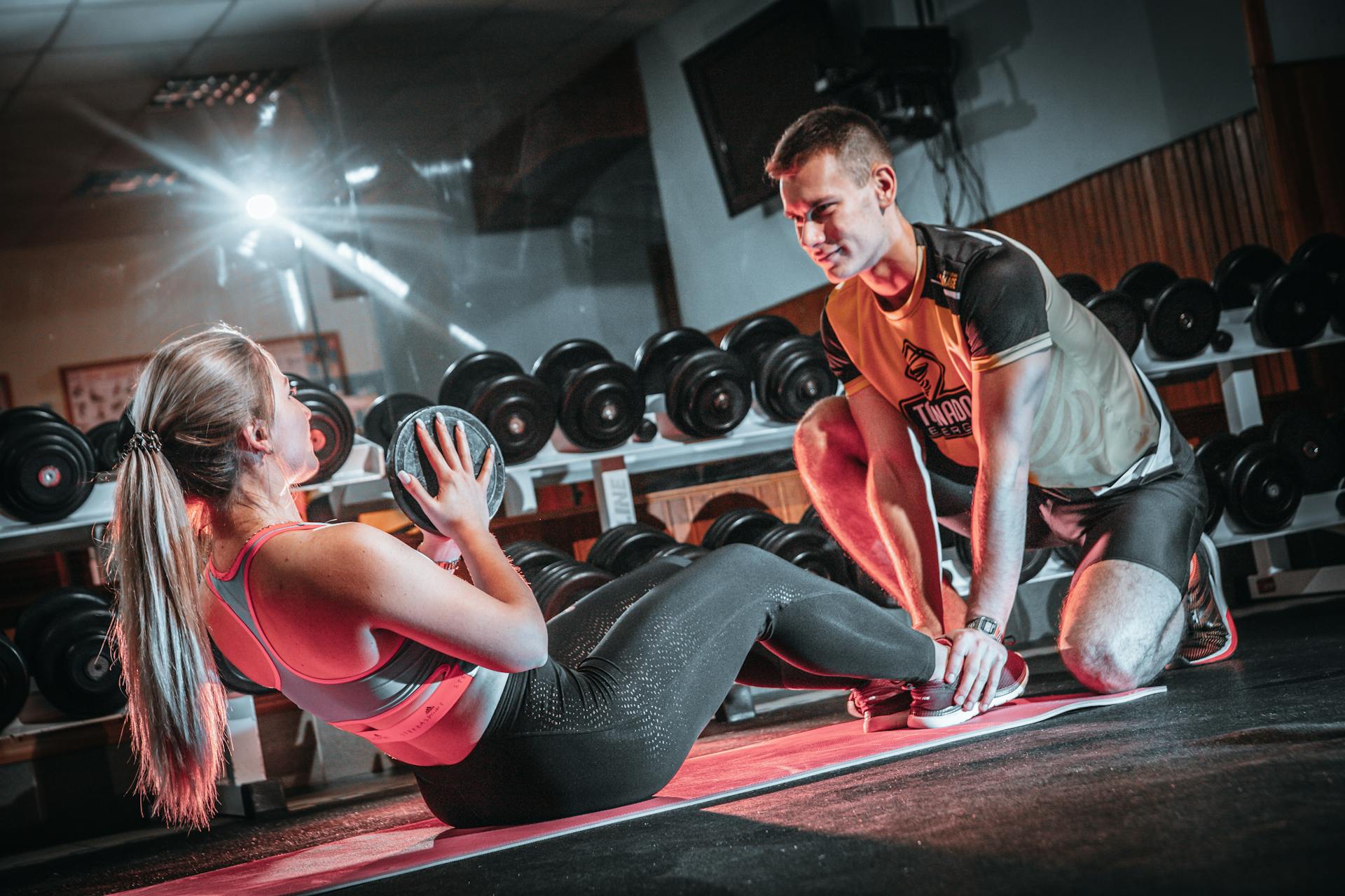 A man in a brown blazer and a woman in a light sweater sit at a desk with a laptop, discussing something. The room is dimly lit, creating a focused atmosphere.