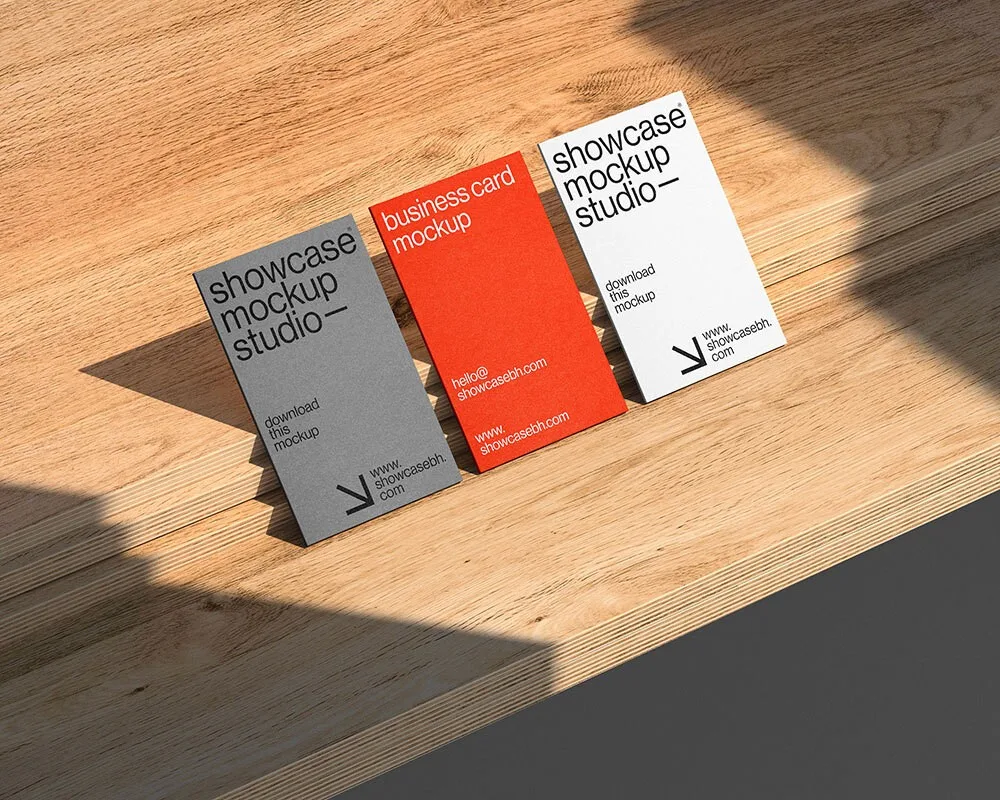 Mockup of three vertically standing business cards in gray, red, and white, placed on a wooden surface. Natural lighting and shadows create a warm and professional presentation ideal for branding.