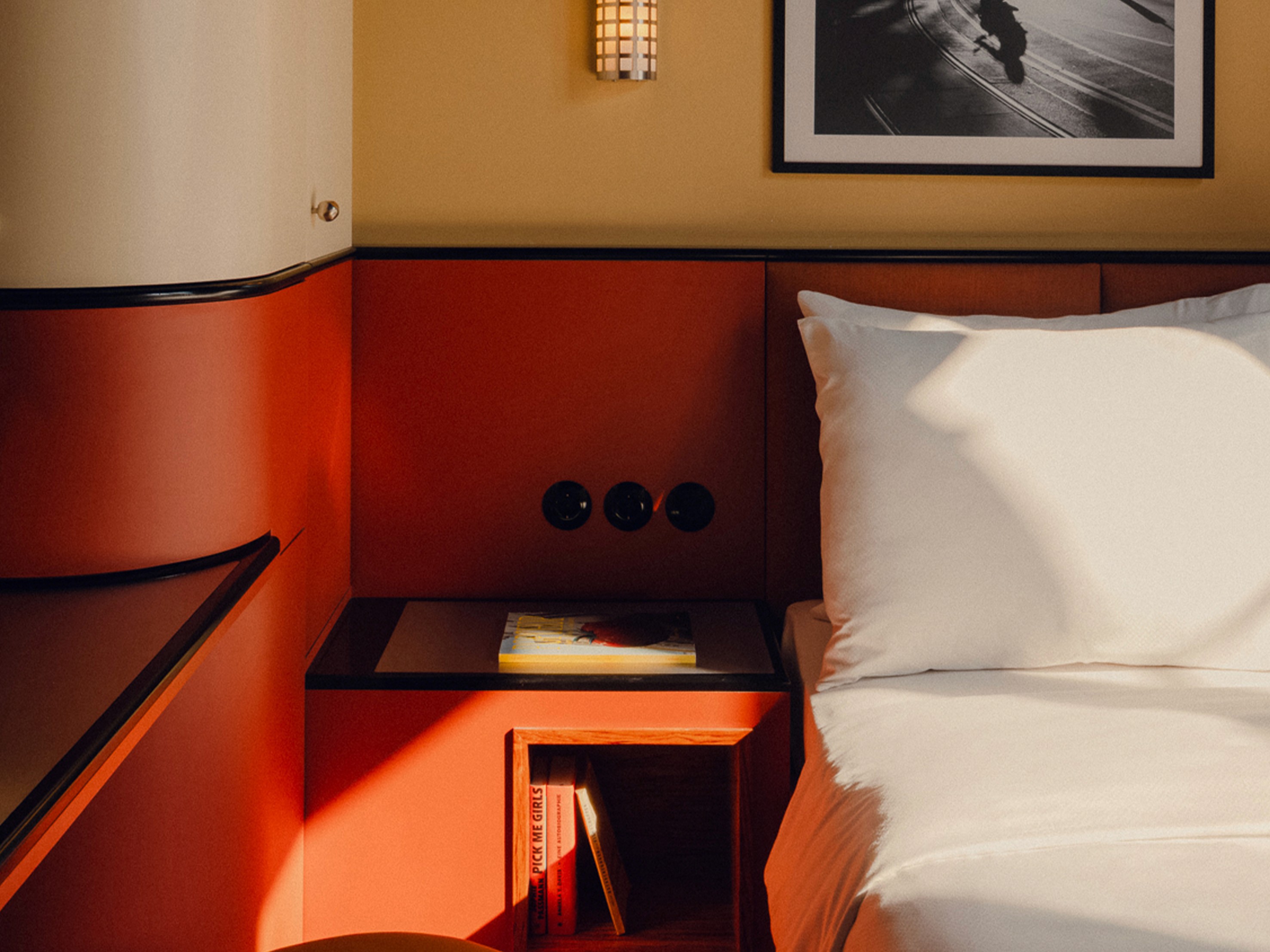 Cozy corner of The Solo room at The Companion Vienna with a red-orange headboard, white bedding, a small wall sconce, and a shelf with a book.