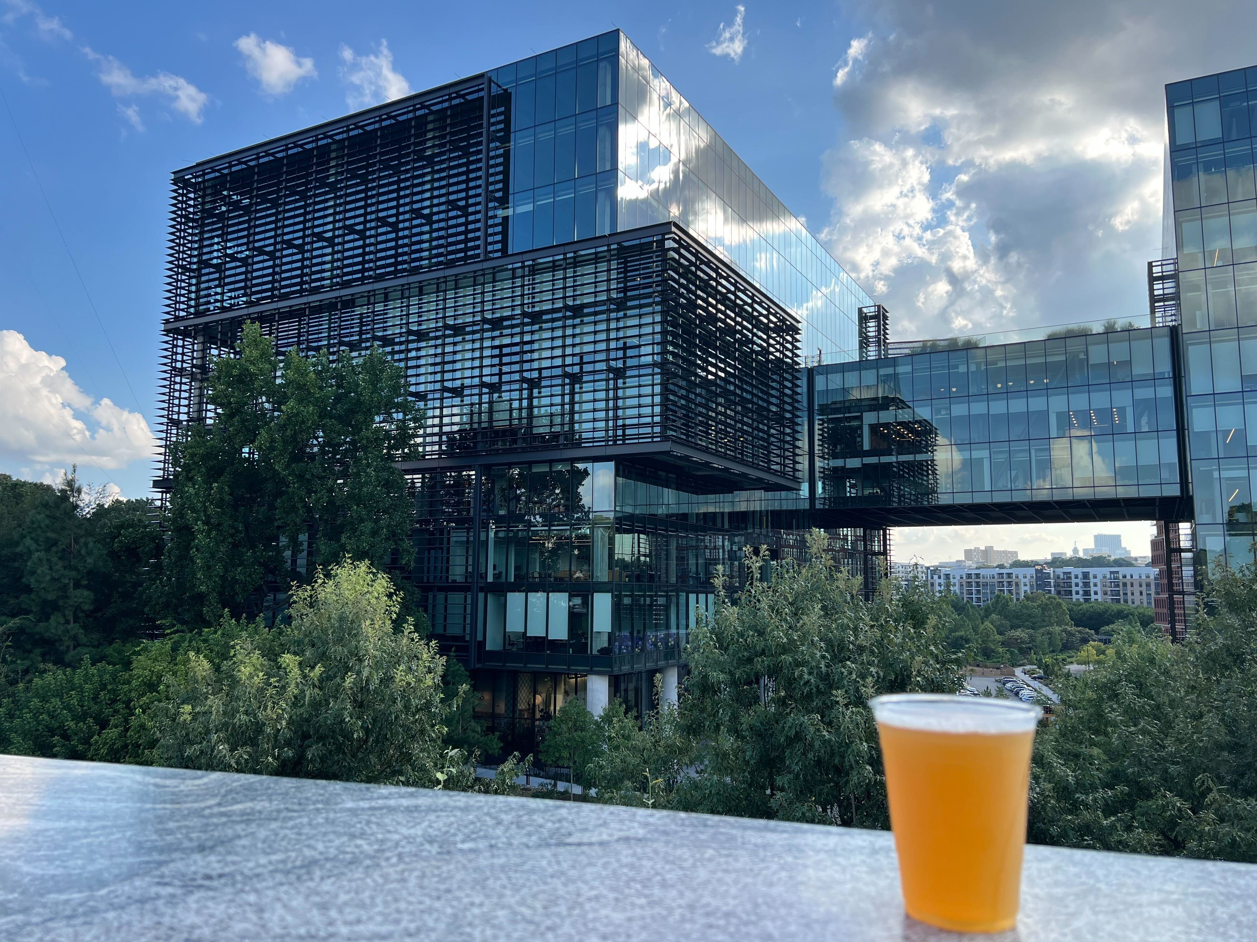 Modern glass office building with skybridge and greenery, viewed from a terrace with a drink in the foreground