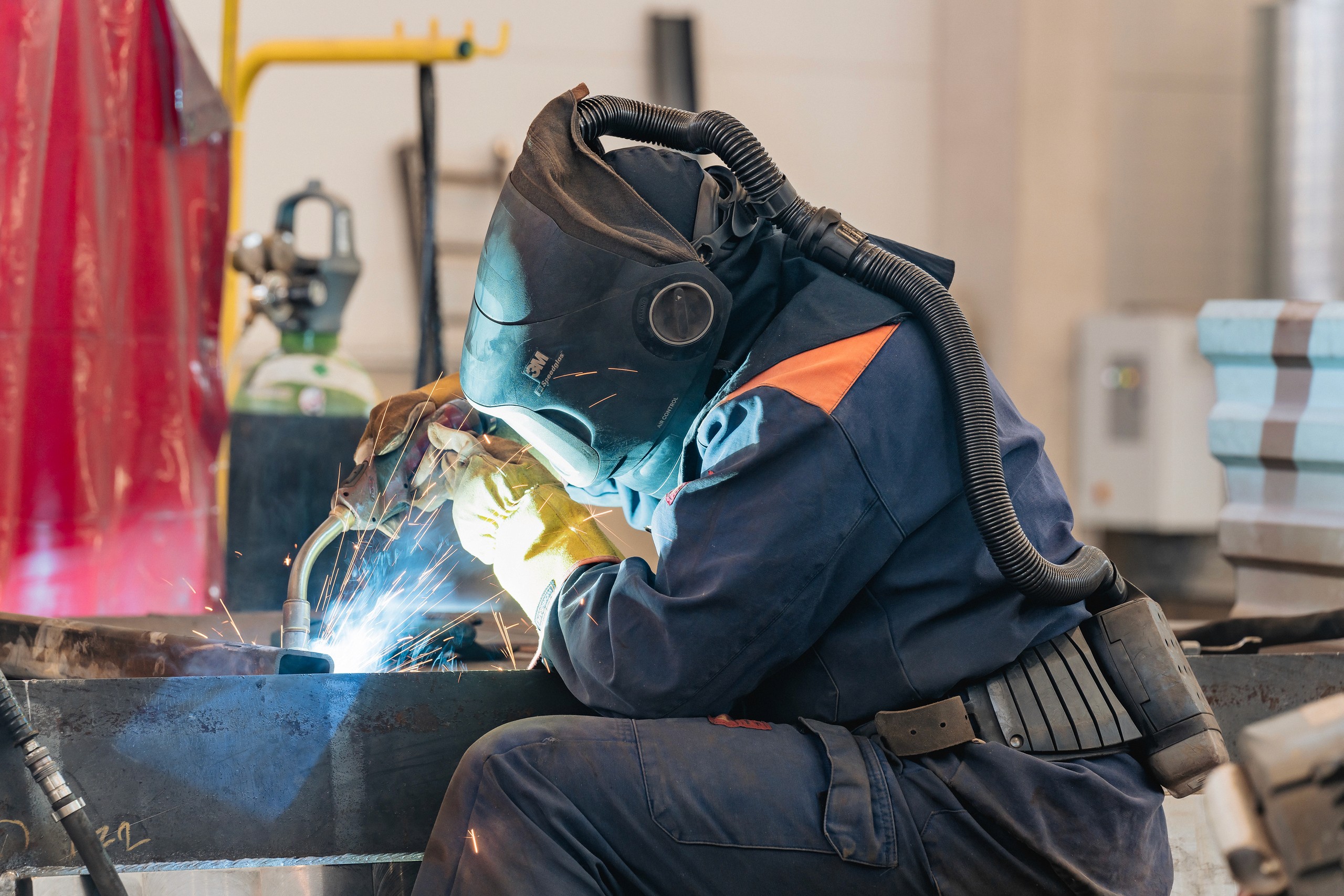 A protected fabricator bends over a steel work surface, with a concentrated burst of welding light.