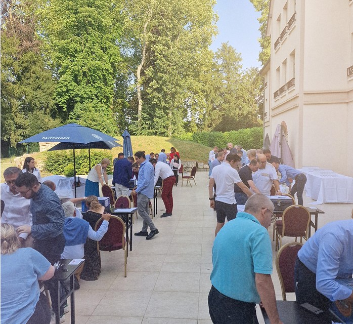 Personnes interagissant autour de tables en extérieur lors d'un événement en plein air, avec un grand bâtiment et des arbres en arrière-plan.