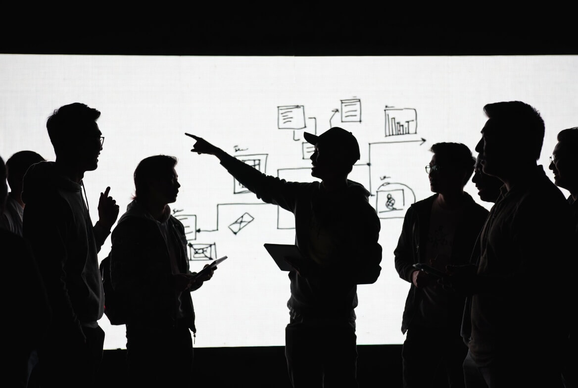 Black and white silhouette of a creative team standing together on stage, actively discussing ideas in front of a brightly lit LED screen. Dynamic group collaboration moment with natural gestures and poses.