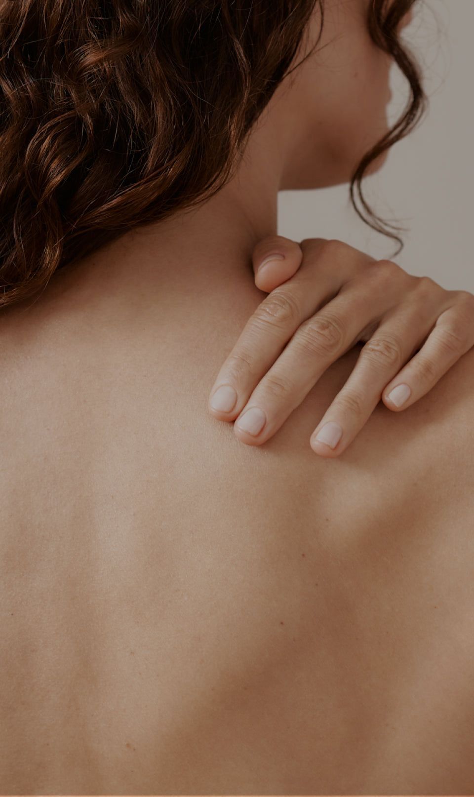 A person with shoulder-length curly hair gently touches their bare shoulder, showcasing smooth skin and a serene, minimalist setting.