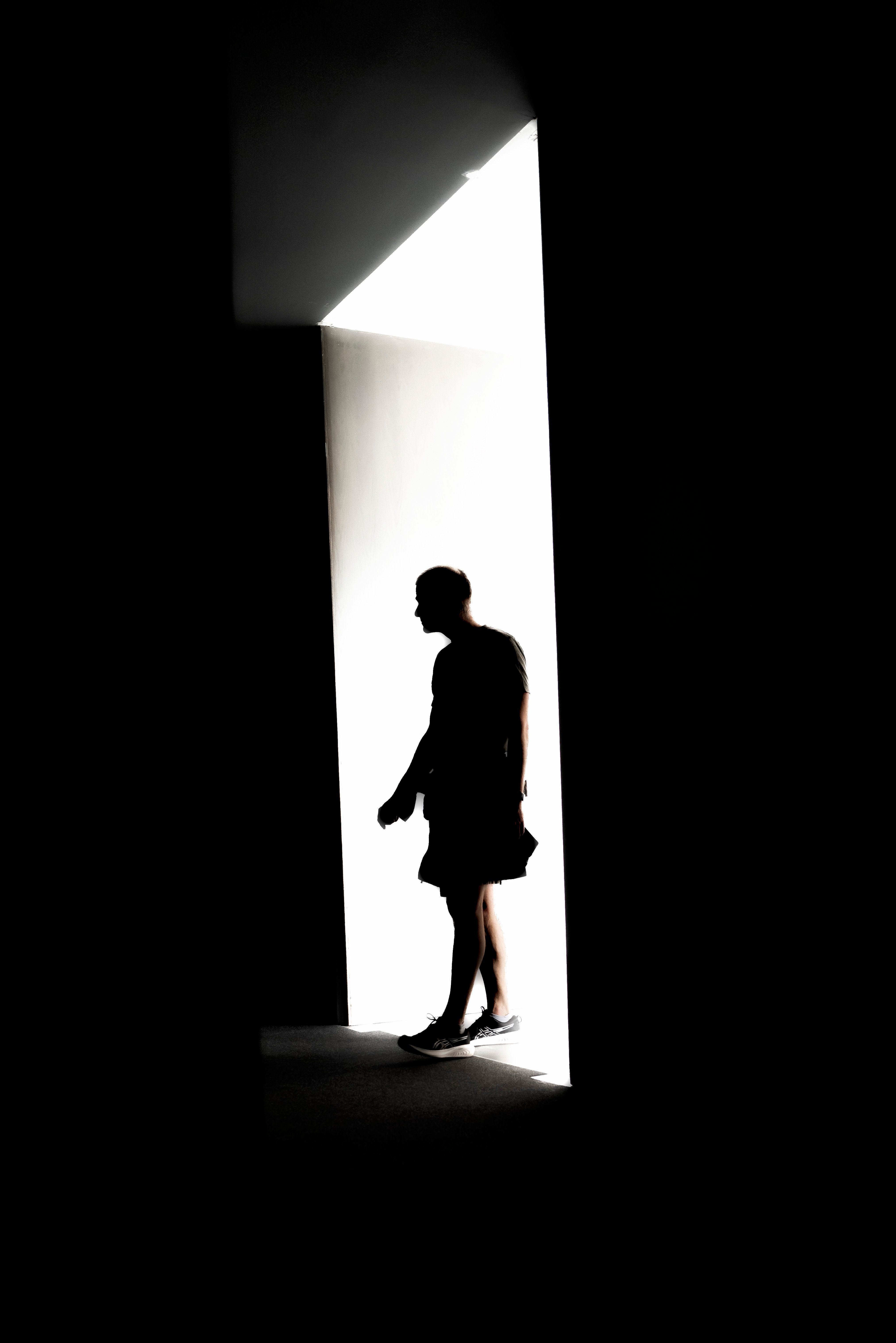 A solitary silhouette walks through a bright, high-contrast doorway, evoking mystery and isolation.