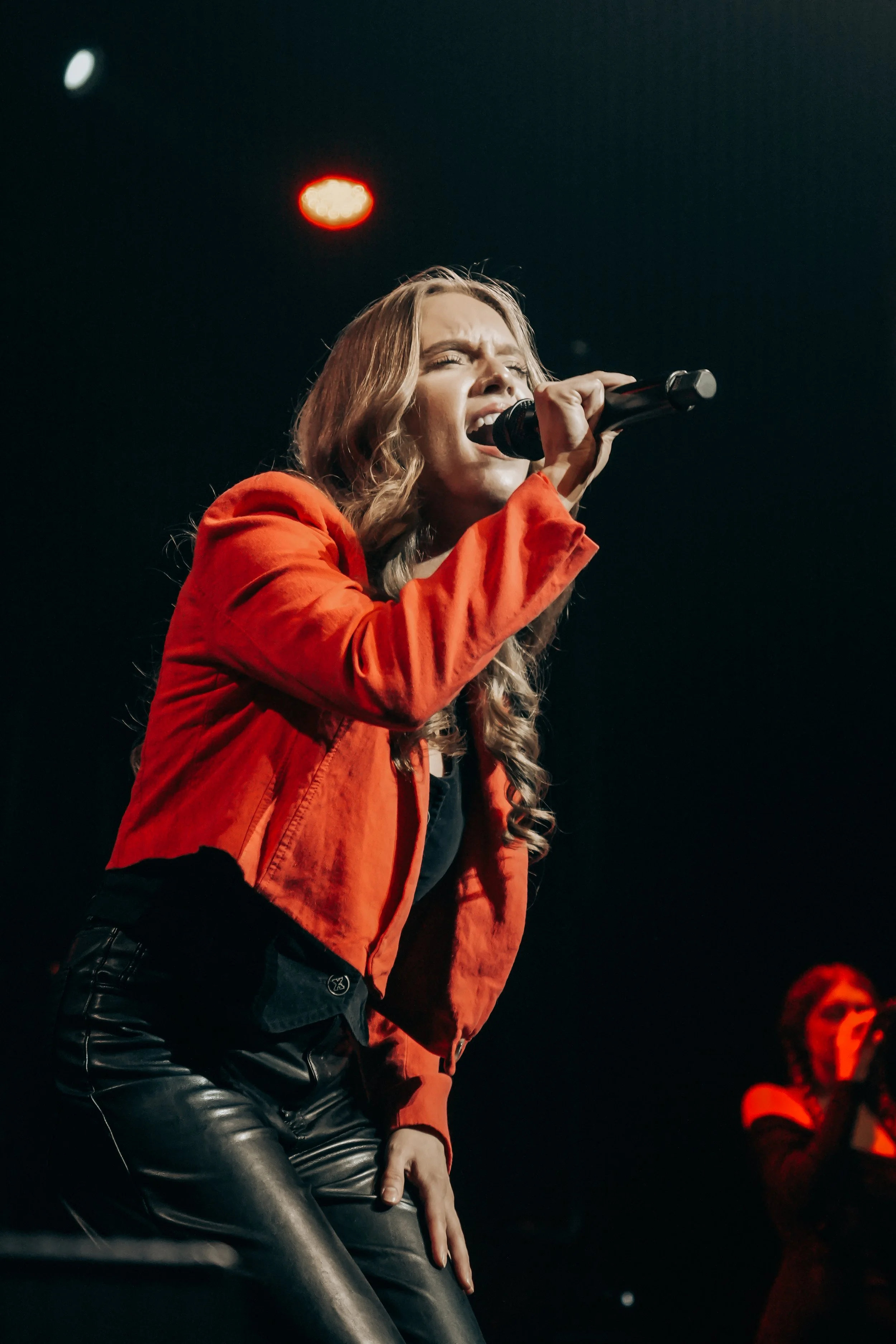 A woman stands beside an old projector, gazing at the camera. The scene is lit with teal and red hues, creating a mysterious and artistic atmosphere.