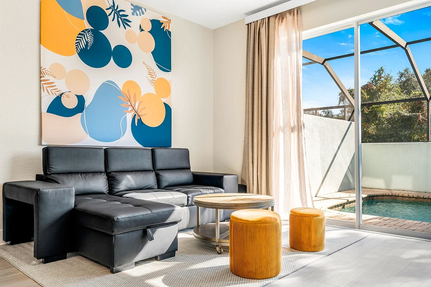Bright Kissimmee Airbnb living room with modern black sectional sofa, colorful wall art, and private splash pool view.
