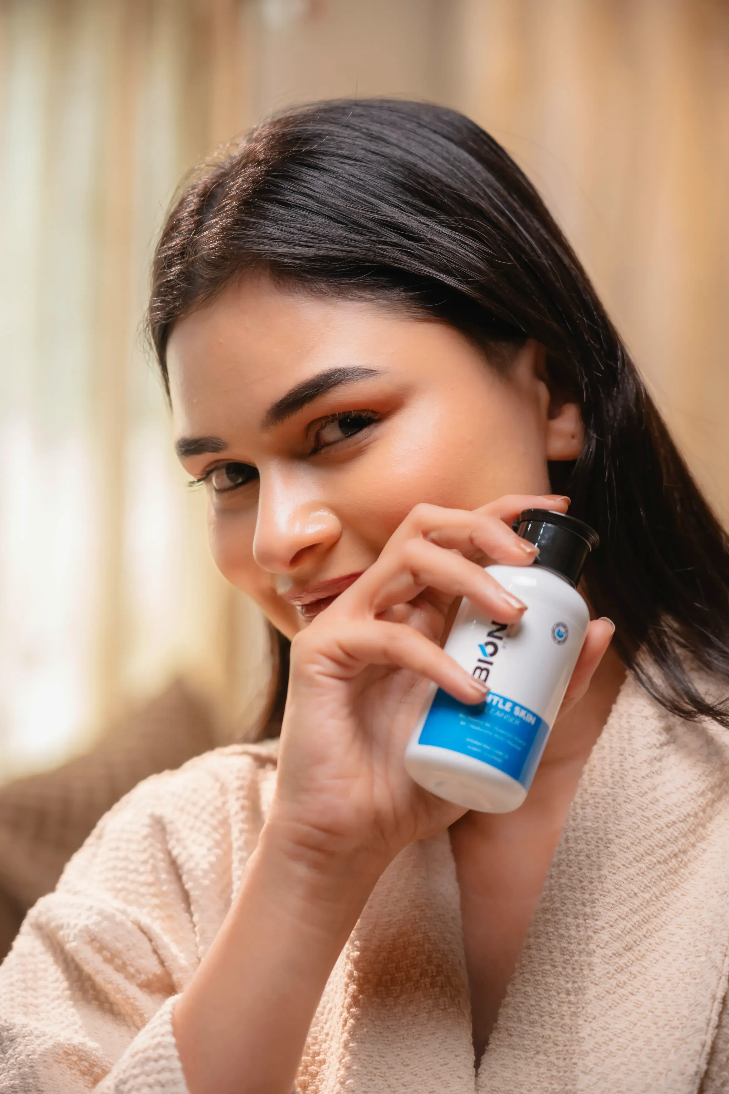 A woman smiles while holding a bottle of pills, sitting in a softly lit room.