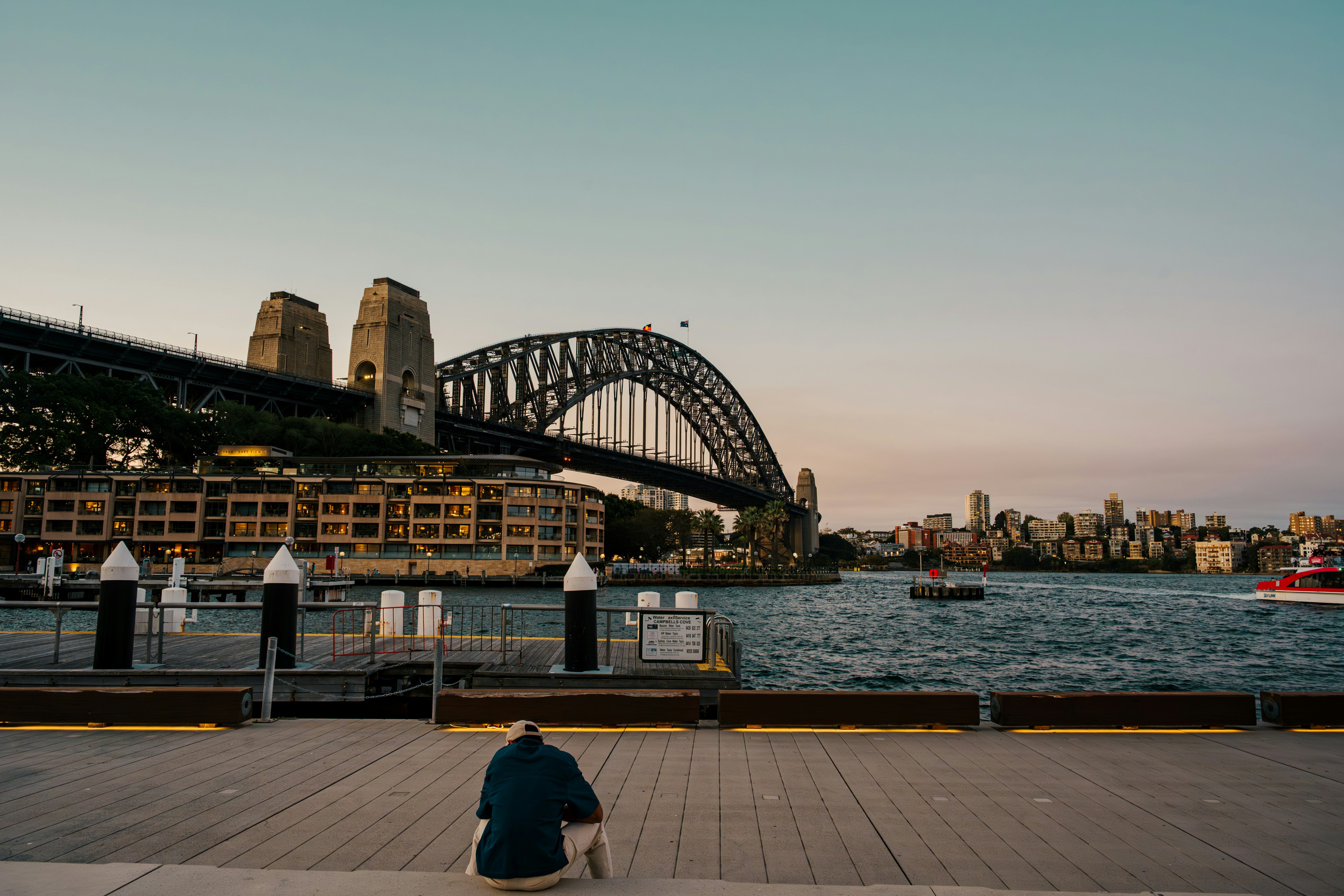 Person sitting by sydney harbour bridge at sunset.