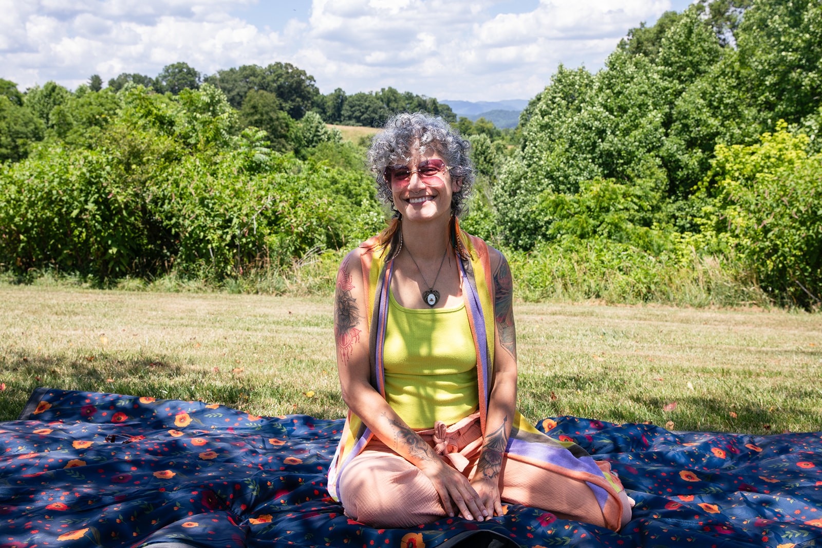 A smiling person sits cross-legged outdoors, surrounded by greenery, wearing a bright yellow top.