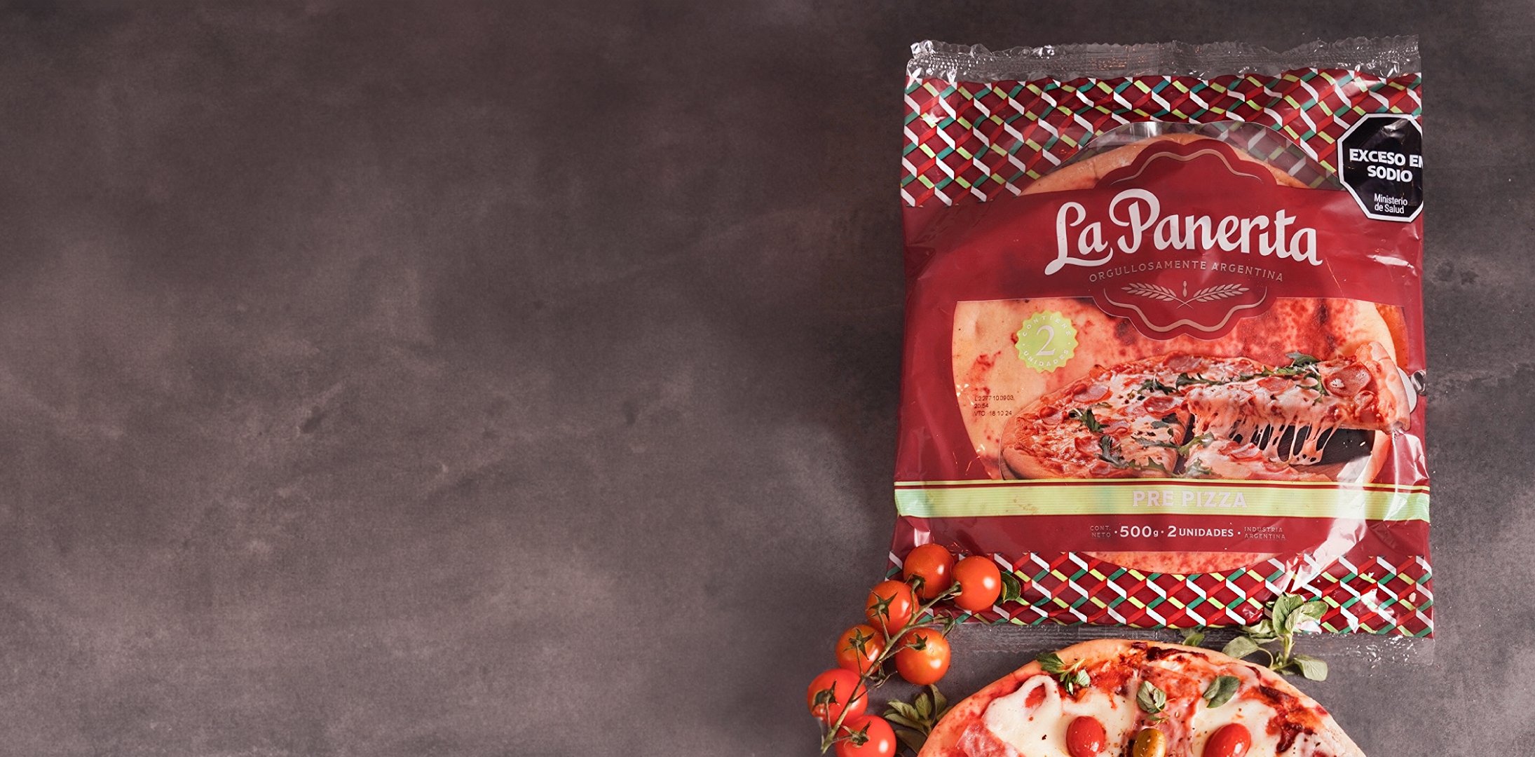 Packaged pizza labeled "La Panerta" on a dark surface, accompanied by fresh vine tomatoes and a partially visible pizza. The mood is appetizing and rustic.