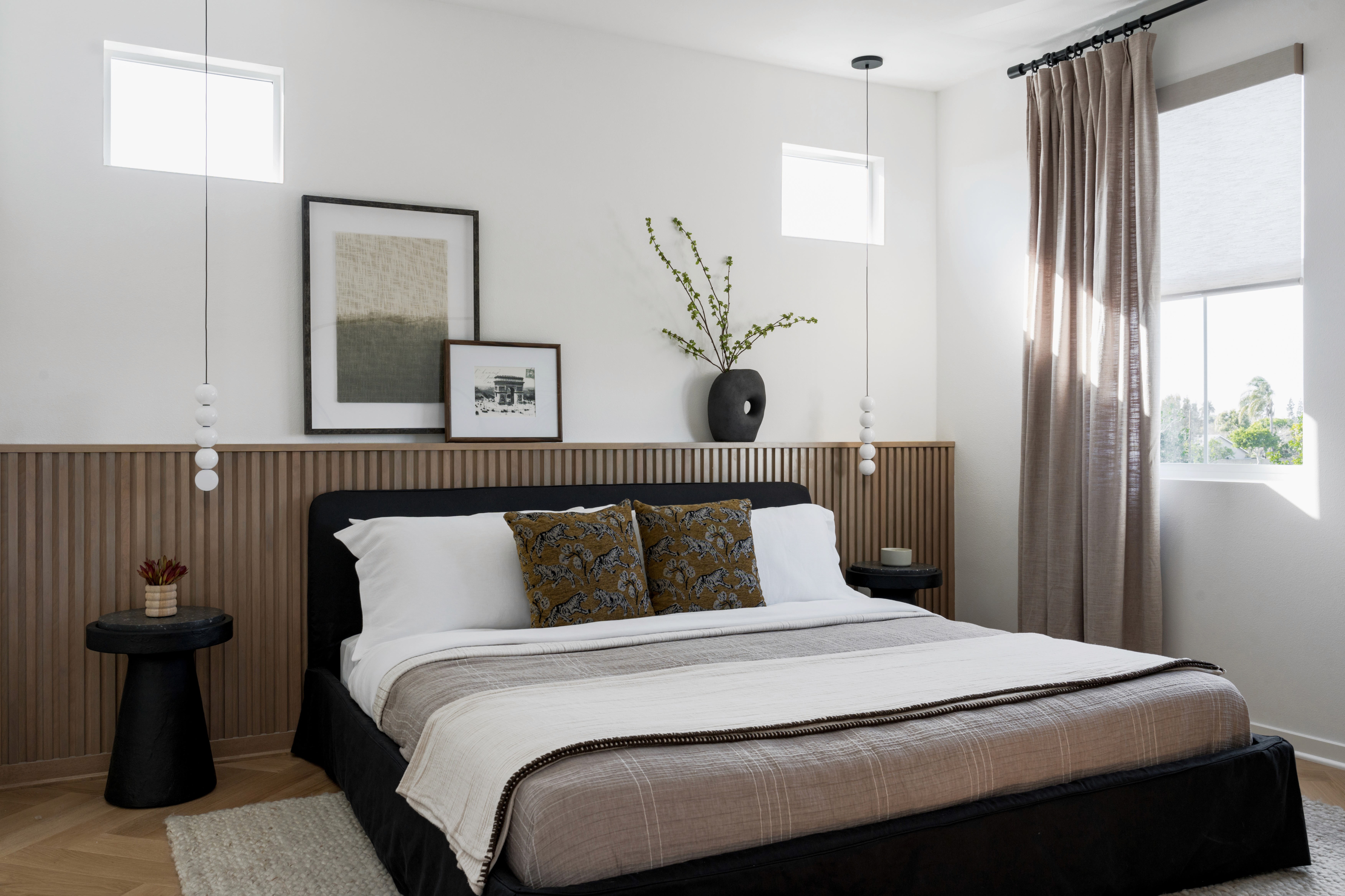 Natural wood fluted wainscoting, artwork above wainscoting, black modern side table, black upholstered bed frame with neutral colored bedding and a variety of layers and textures, large open windows with floor to ceiling drapers, eclectic bulb pendant lighting