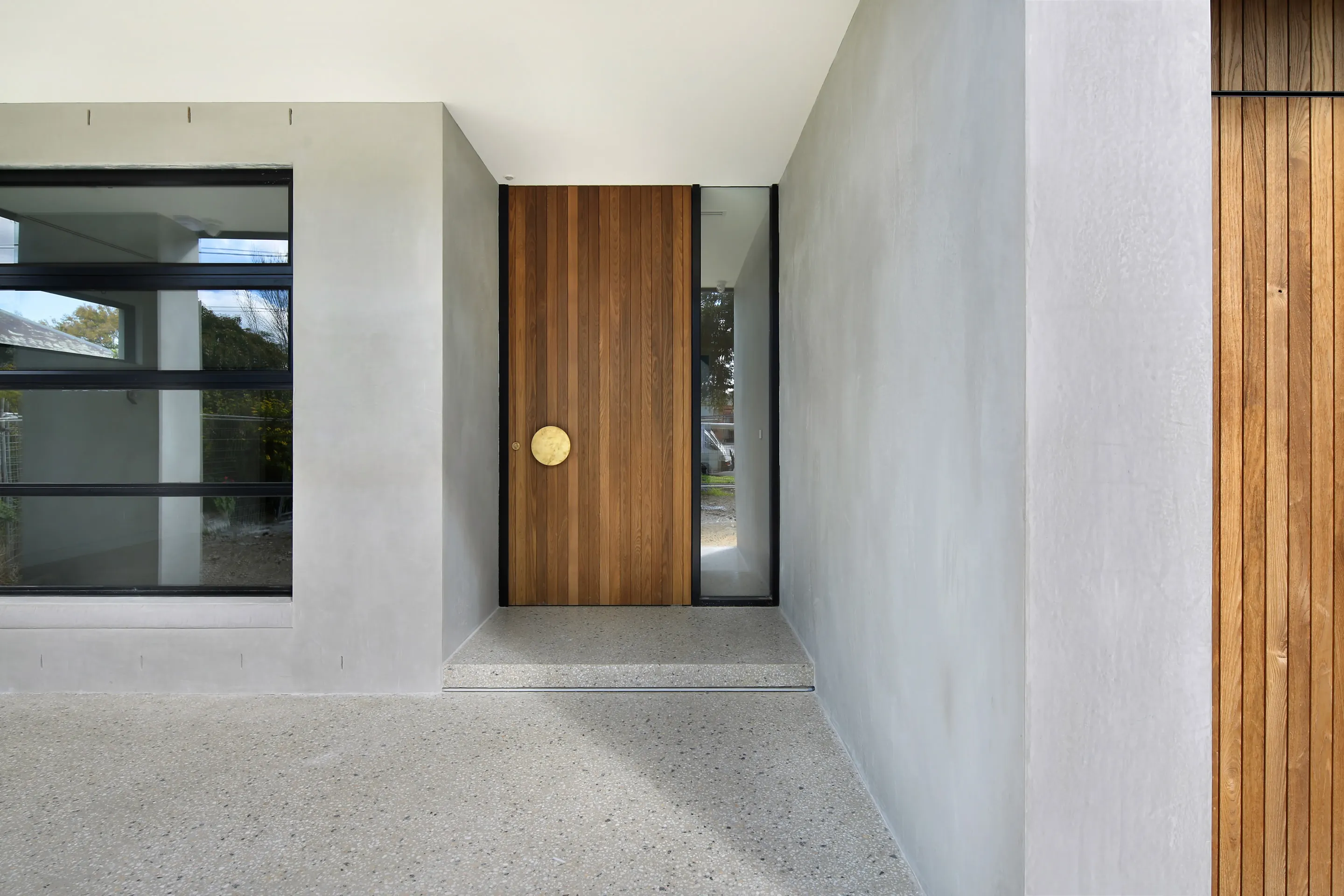 Covered entry porch with rendered walls, polished-concrete floor, a black-framed picture window and a timber-batten pivot door with a round brass handle