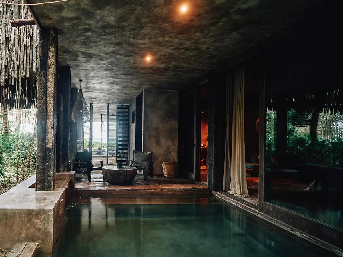Premier Suite en Nômade Tulum con gran ventana de cristal con vista a la piscina privada y las palmeras, y cómoda área de asientos con sofá y mesa central.