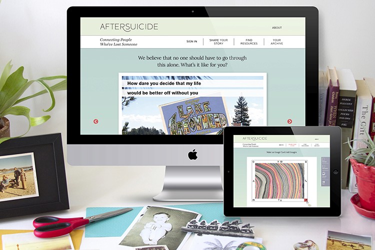 A desktop computer monitor displays a webpage titled "AFTERSUICIDE" with the tagline "Connecting People Who've Lost Someone." The webpage includes a quote: "We believe that no one should have to go through this alone. What's it like for you?" Below the quote, a colorful graphic is visible. A tablet screen shows the same webpage with a colorful, circular design. To the left of the computer, a pair of scissors rests on a light blue cutting mat next to a framed photo and a small potted plant. To the right of the computer, a stack of books and a small potted plant are visible.