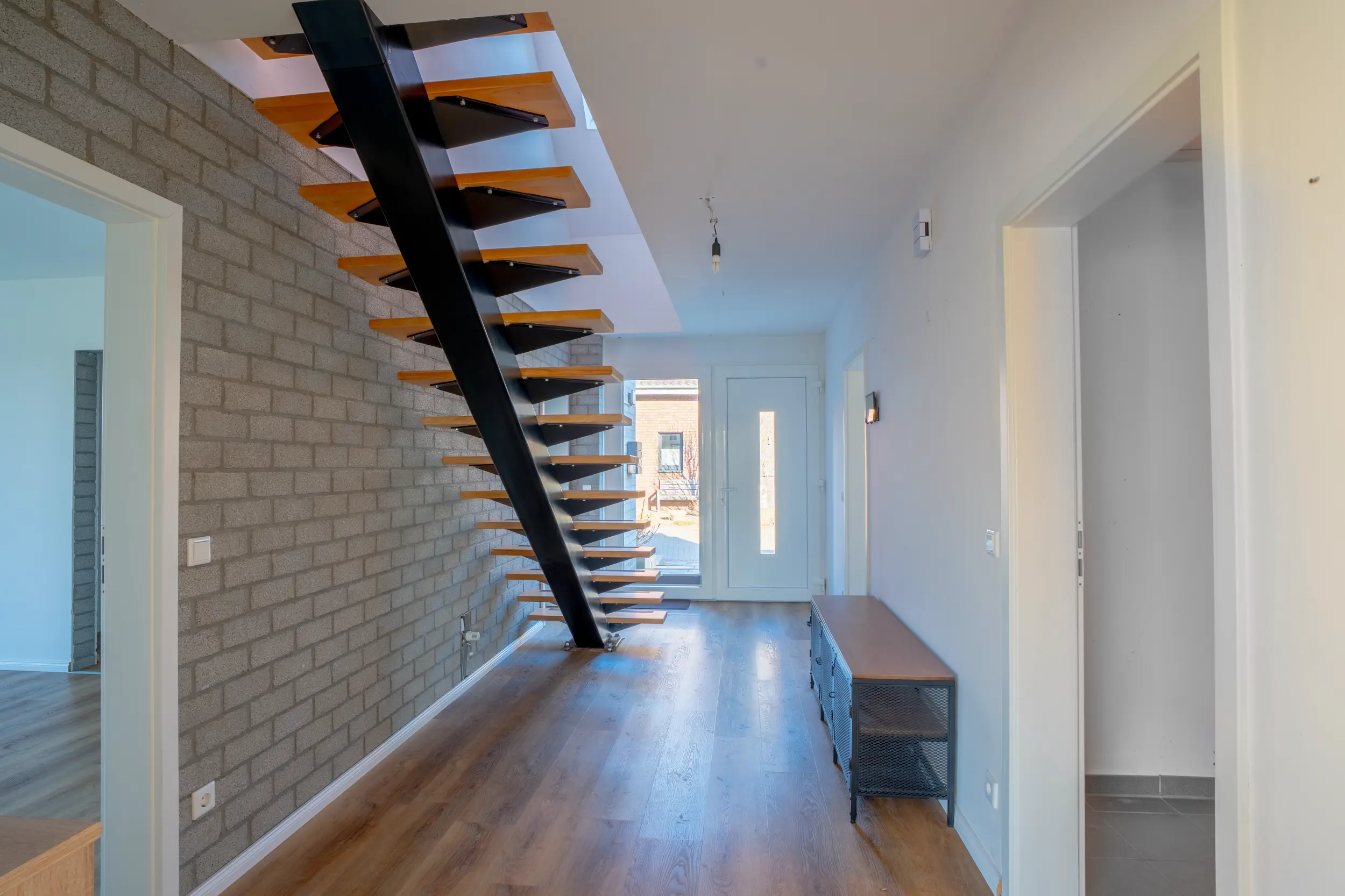 Eingangsbereich mit freitragender Treppe aus Holzstufen und Stahlwange an grauer Klinkerwand, Eichenholzboden und Haustür mit Glasausschnitt