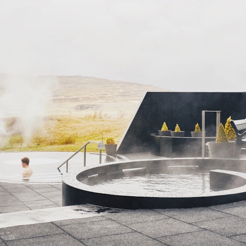 Person sitting in a steaming outdoor hot tub with a mountainous landscape in the background and modern design elements around.