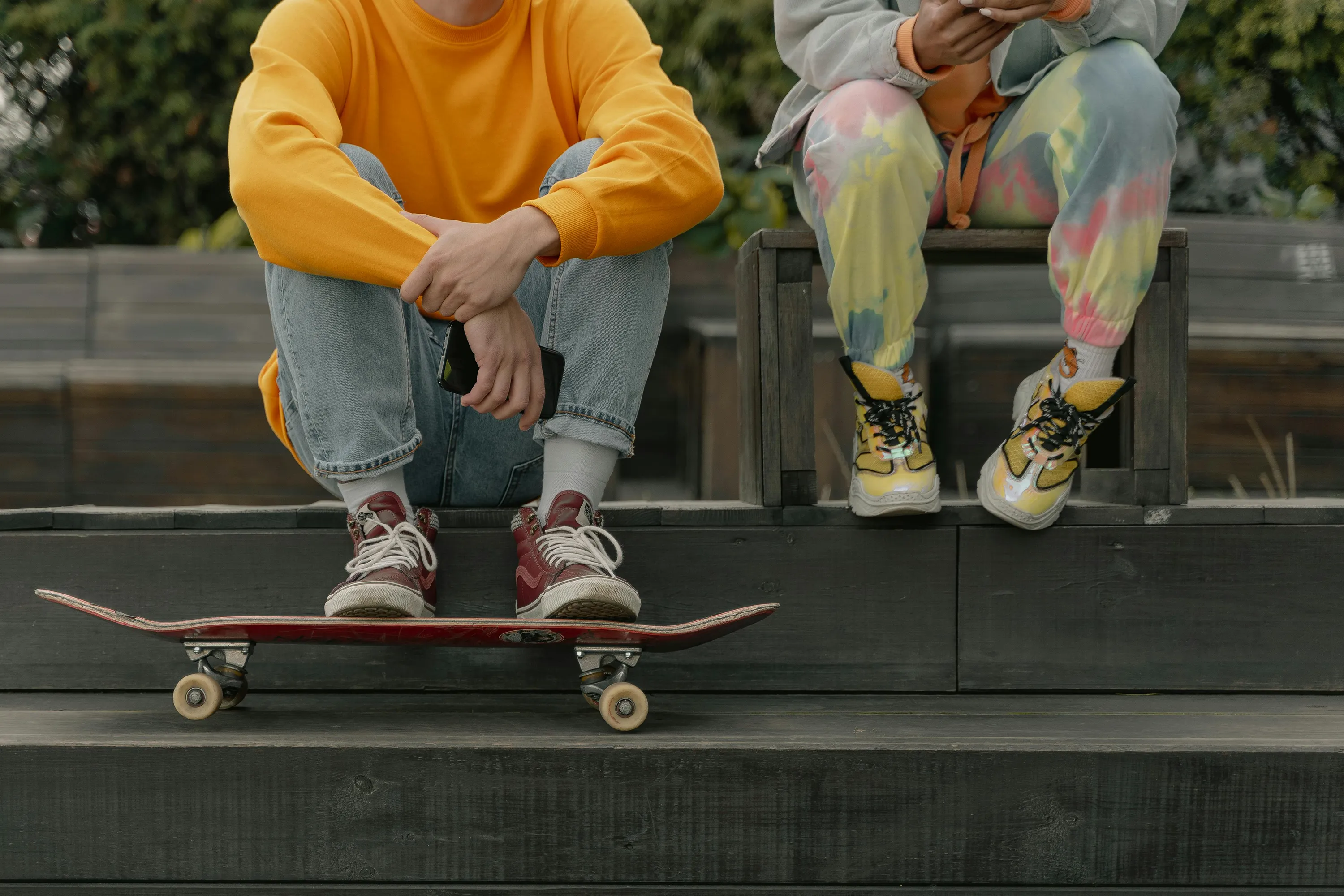 The lower half of two people sitting together, one with their feet on a skateboard