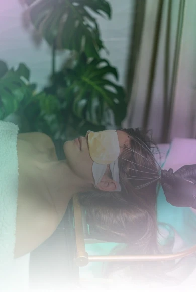 A person lies back on a spa chair with eyes covered by a soothing gel mask, surrounded by lush green plants and soft lighting, creating a tranquil and relaxing spa environment.