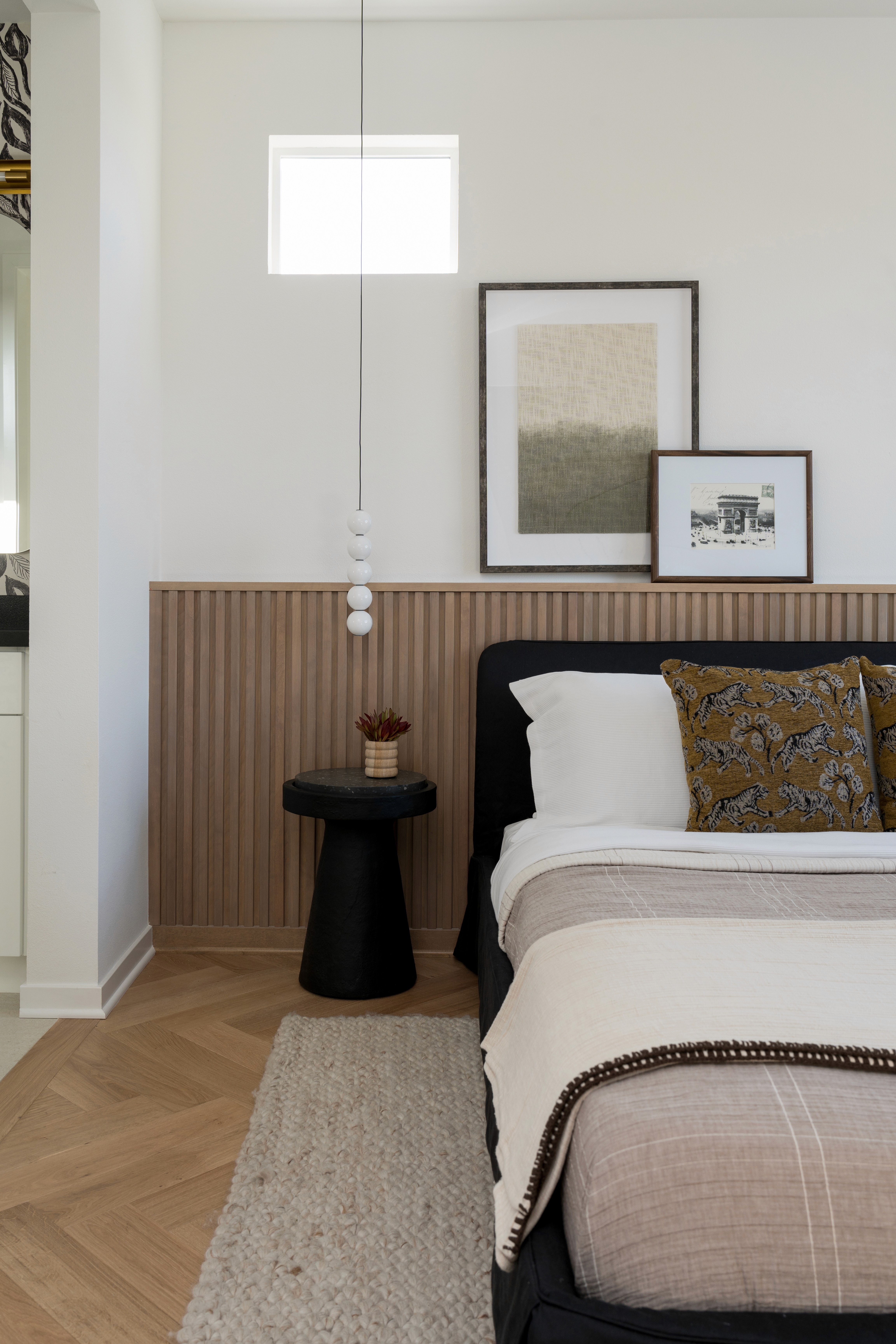 Natural wood fluted wainscoting, artwork above wainscoting, black modern side table, black upholstered bed frame with neutral colored bedding and a variety of layers and textures