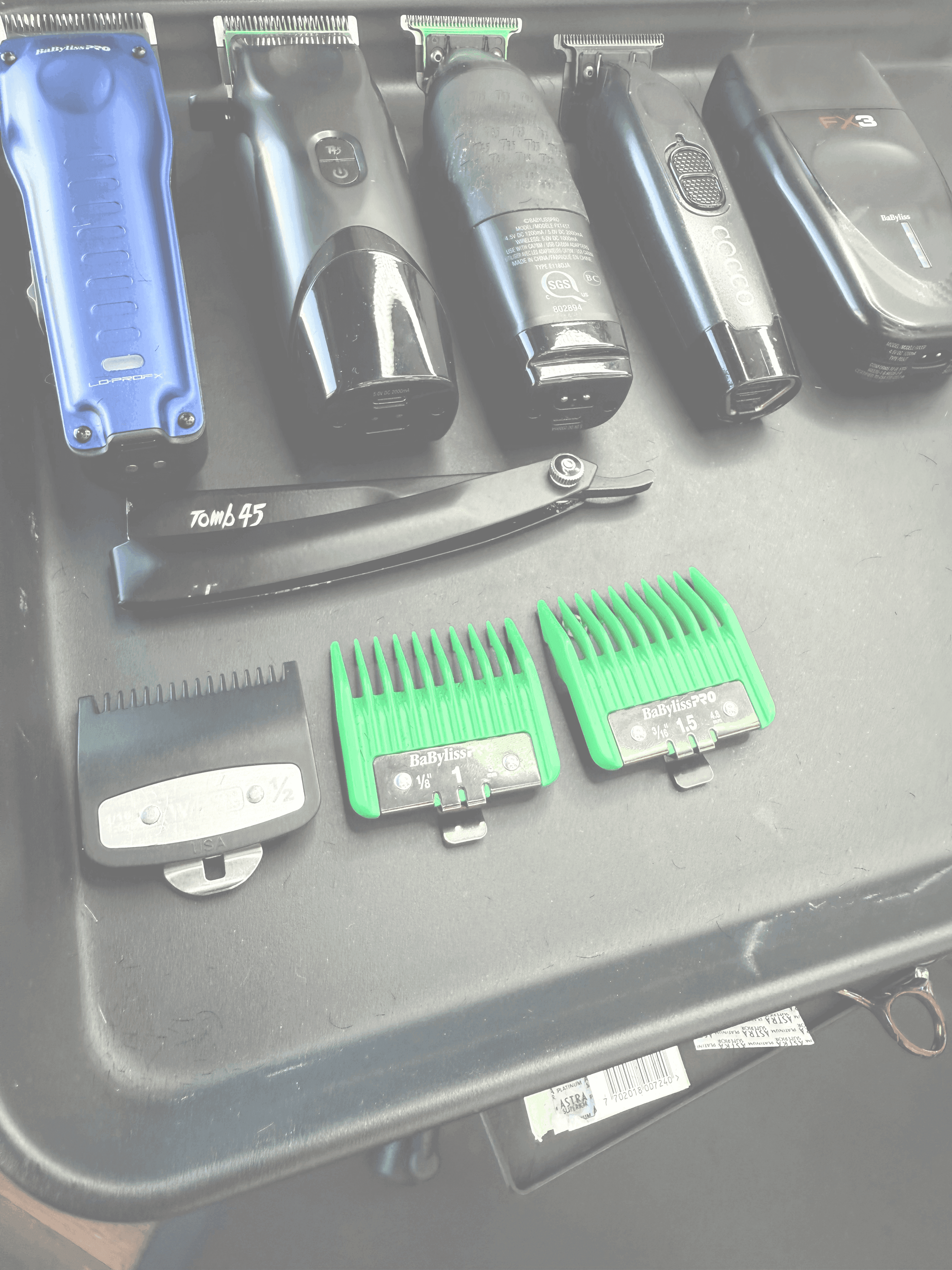 Close-up of barber tools on a workstation, including clippers, trimmers, a straight razor, green clipper guards, and other grooming accessories.