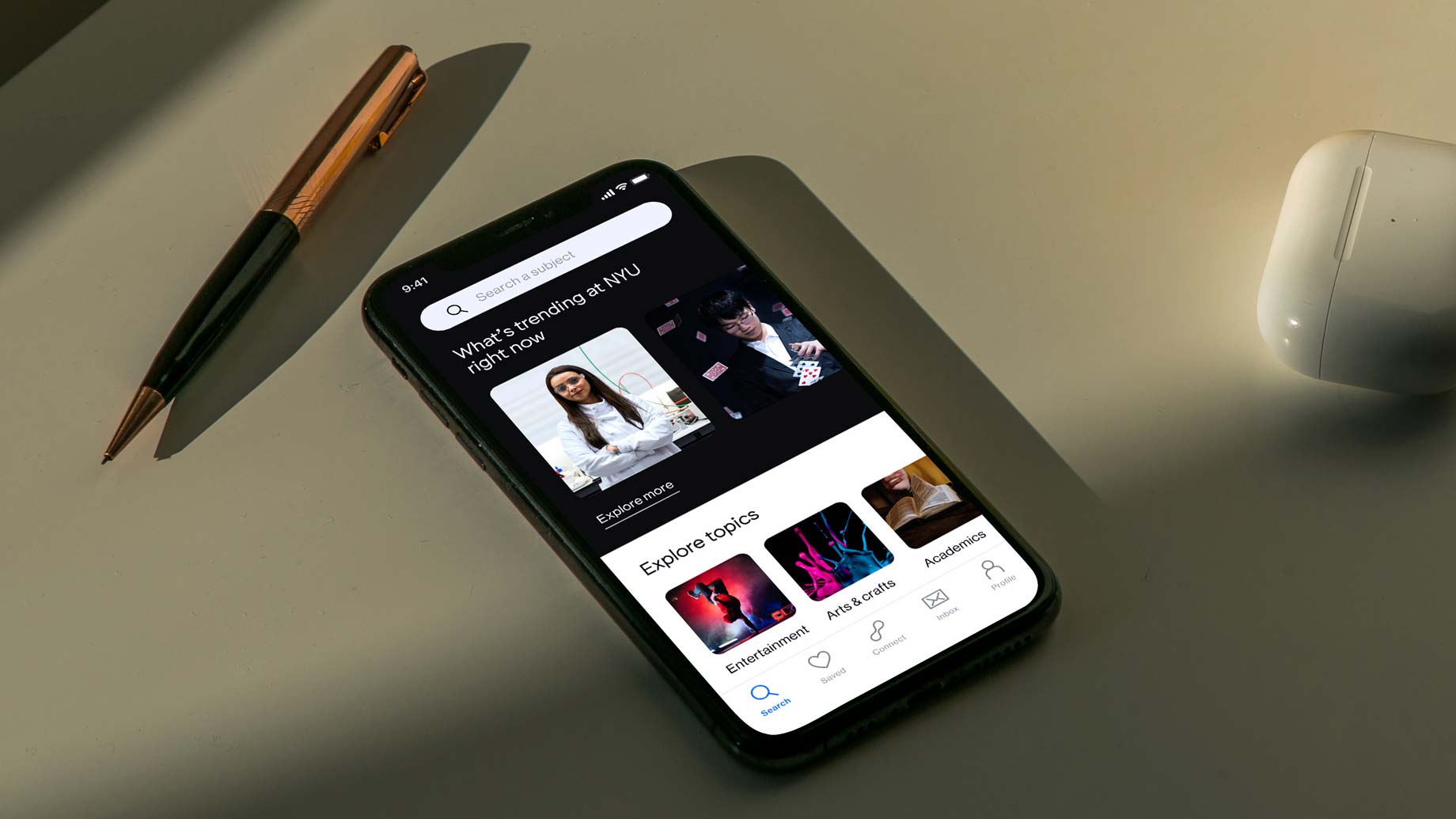 A black smartphone lies on a light-colored surface, angled slightly to the right. The screen displays a social media interface showing a post titled “What’s trending at NFT?” with a photo of a woman in a white shirt and glasses. Below the post are tabs labeled “Explore topics,” “Entertainment,” “Academics,” and “Arts & Crafts.” The time displayed on the phone is 9:41. A gold pen and a white wireless mouse are positioned to the left of the phone.