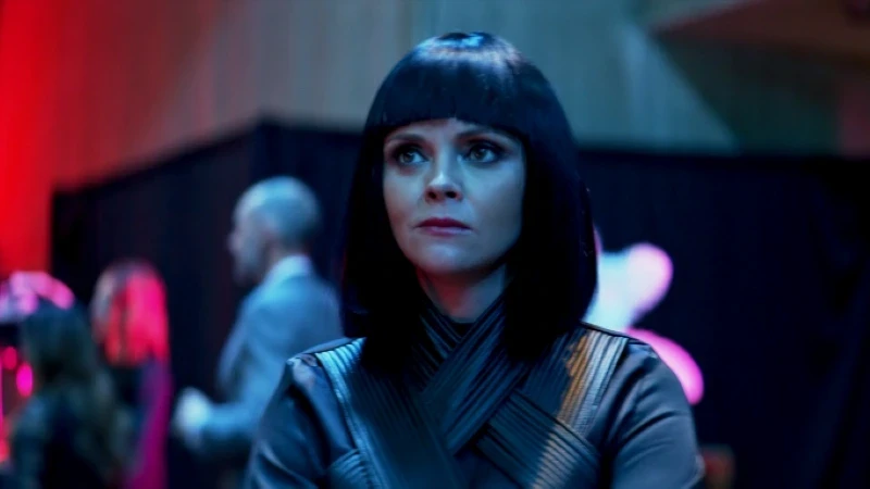 A woman with straight, shoulder-length black hair and bangs wears a dark-coloured, textured outfit, standing in a dimly lit room with out-of-focus people and vibrant red and blue lighting in the background.