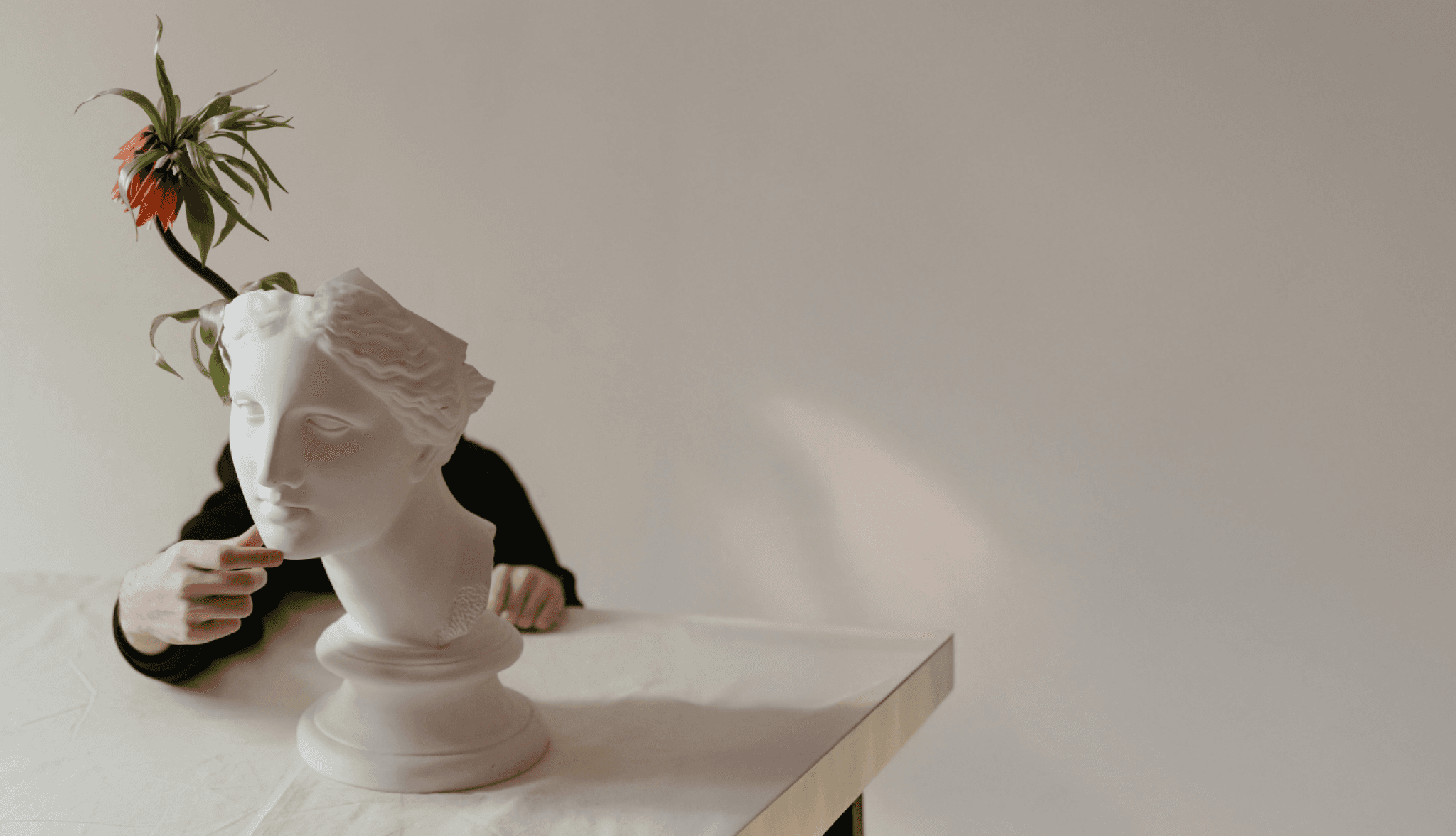 A white plaster bust sculpture with an orange flower placed on top, partially obscured by a hand, creating a minimalist and contemplative visual composition