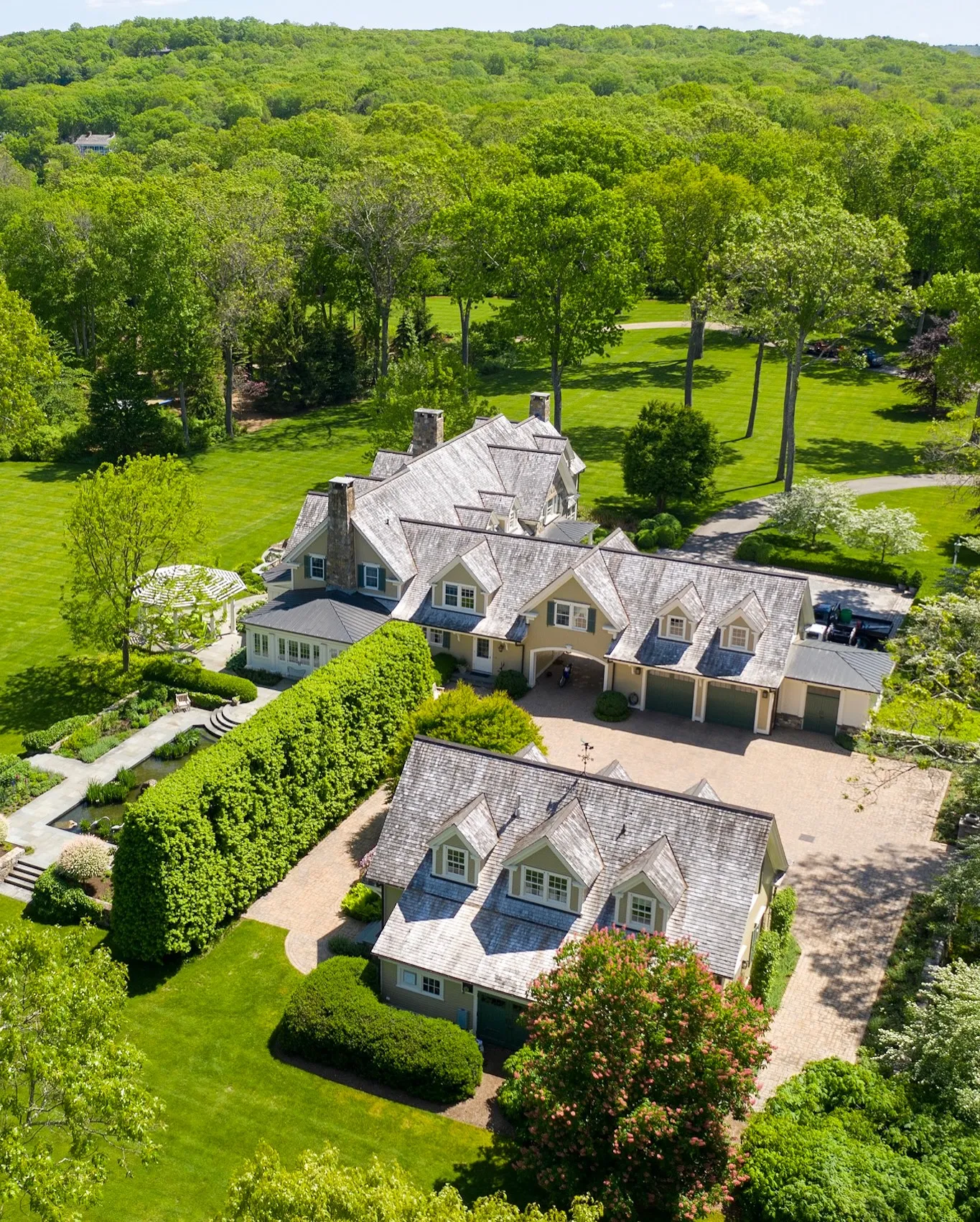 Aerial view of a sprawling estate with a large white house featuring steeply pitched roofs, multiple chimneys, and dormer windows, surrounded by lush green lawns, tall trees, and a circular driveway.