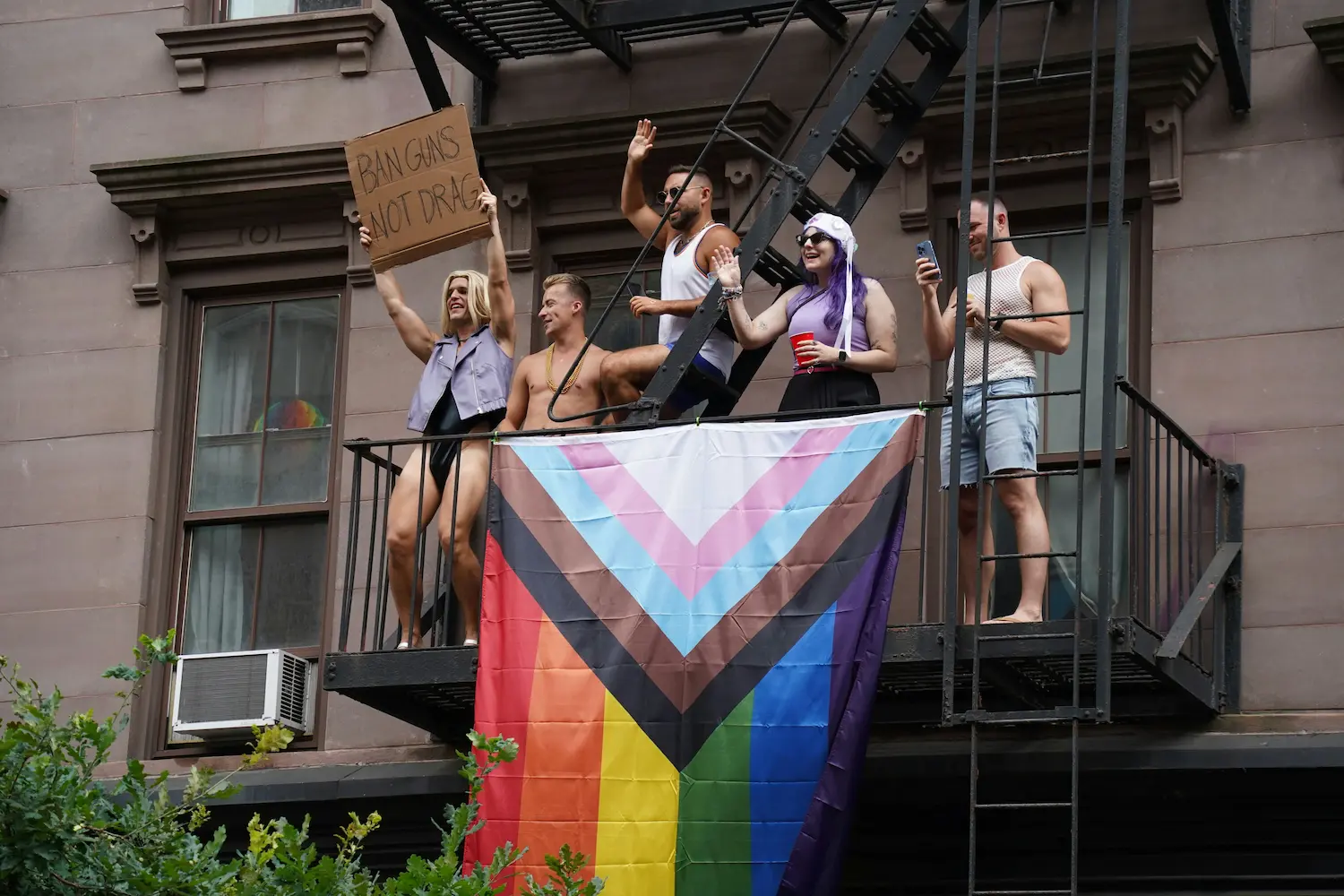 LGBTQ+ community members displaying a Progress Pride flag and protest sign from a fire escape during a public demonstration, symbolizing queer activism, solidarity, and collective resilience.