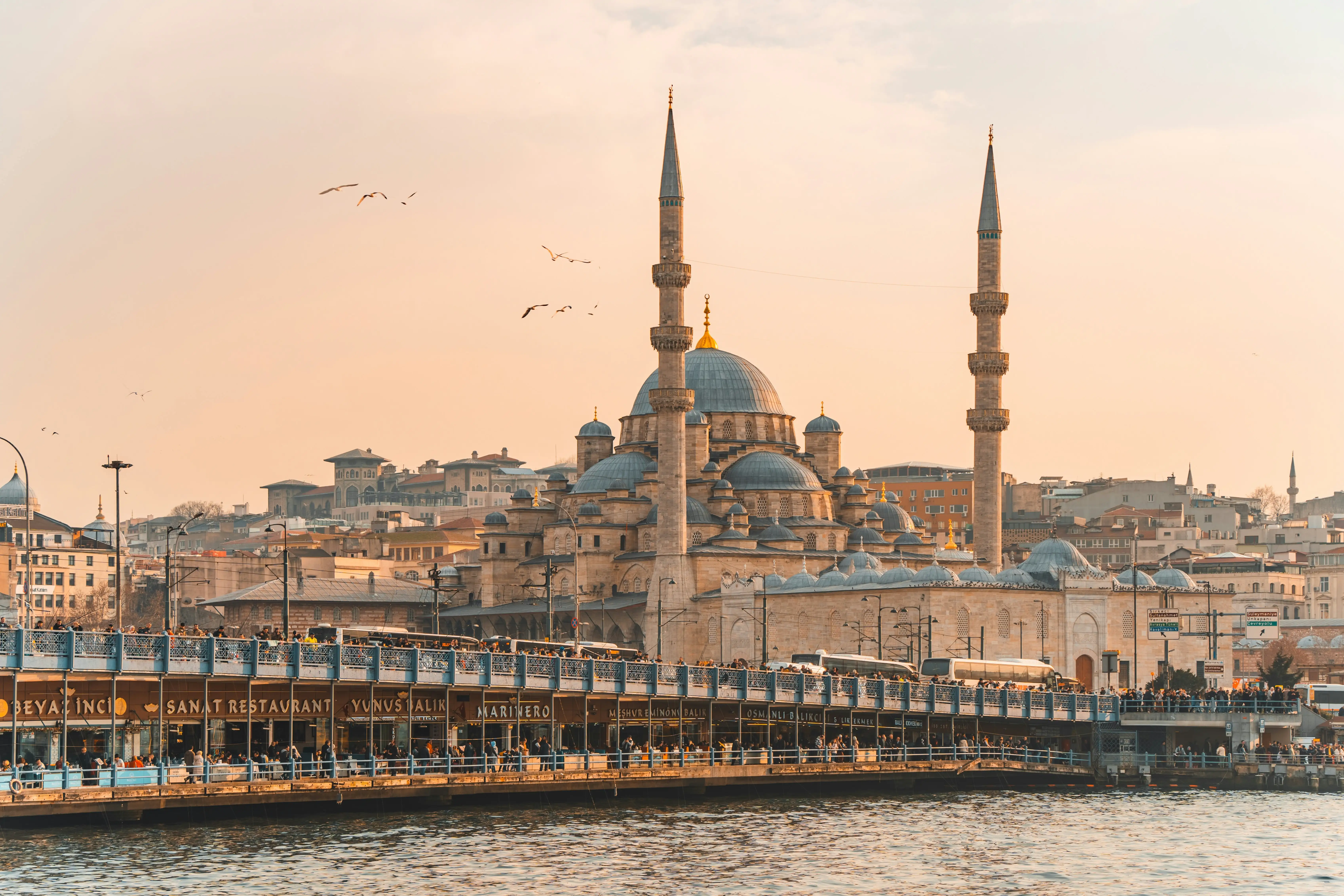 A large historic mosque with two tall minarets, set against a soft, hazy skyline and reflecting in the water below.