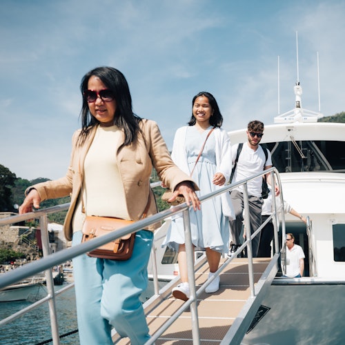 Tři lidé sestupují po lávce z lodi za slunečného dne. Žena vpředu má na sobě sluneční brýle a béžovou bundu.