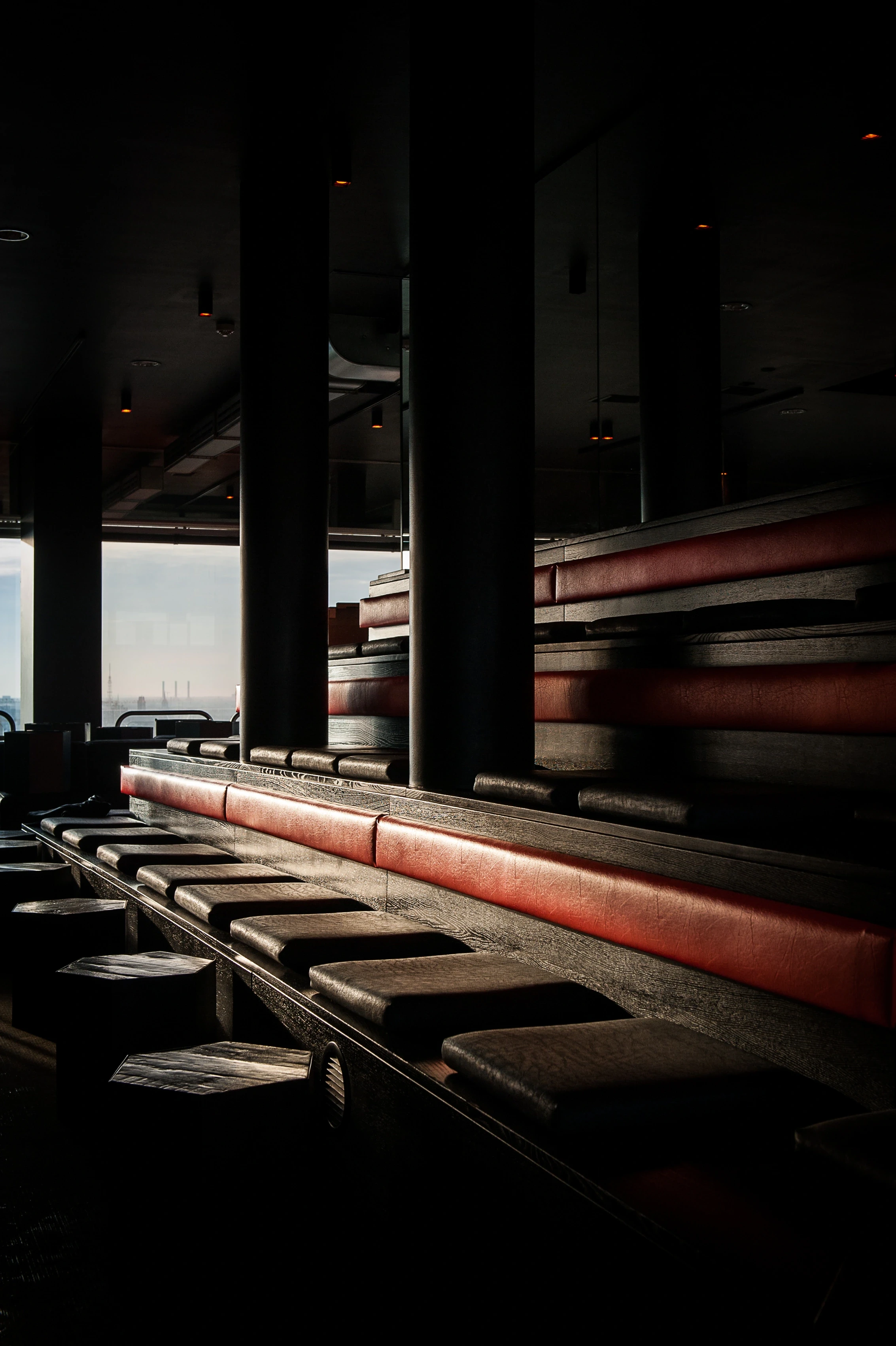 Tiered seating at SOLAR SKY LOUNGE with dark leather cushions and red upholstered backrests catching warm light, black columns and a city view through the window.