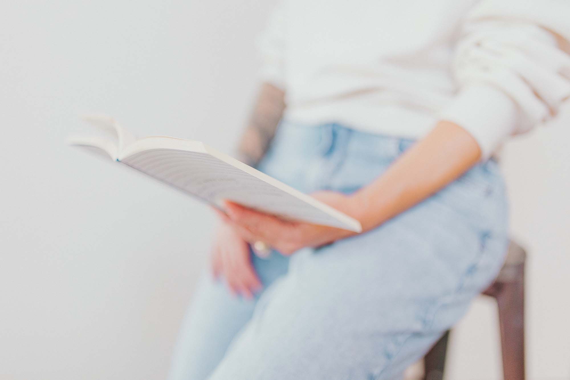 A white woman wearing a white sweatshirt and a light pair of jeans sitting on a stool holding an open white book in her hands