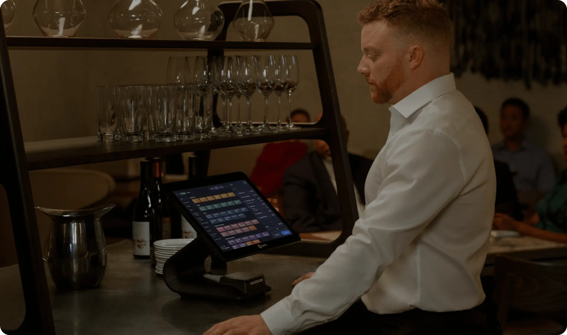 A person in a white shirt is standing at a modern touchscreen point-of-sale system in a dimly lit restaurant, with wine glasses and bottles visible in the background, embodying professional service in an upscale dining setting in Bradenton, FL, showcasing SpotOn technology.