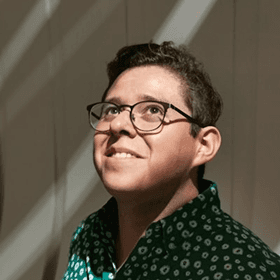 A person wearing glasses and a green shirt with a white pattern looks upwards, bathed in soft, filtered light, creating a serene and contemplative atmosphere.