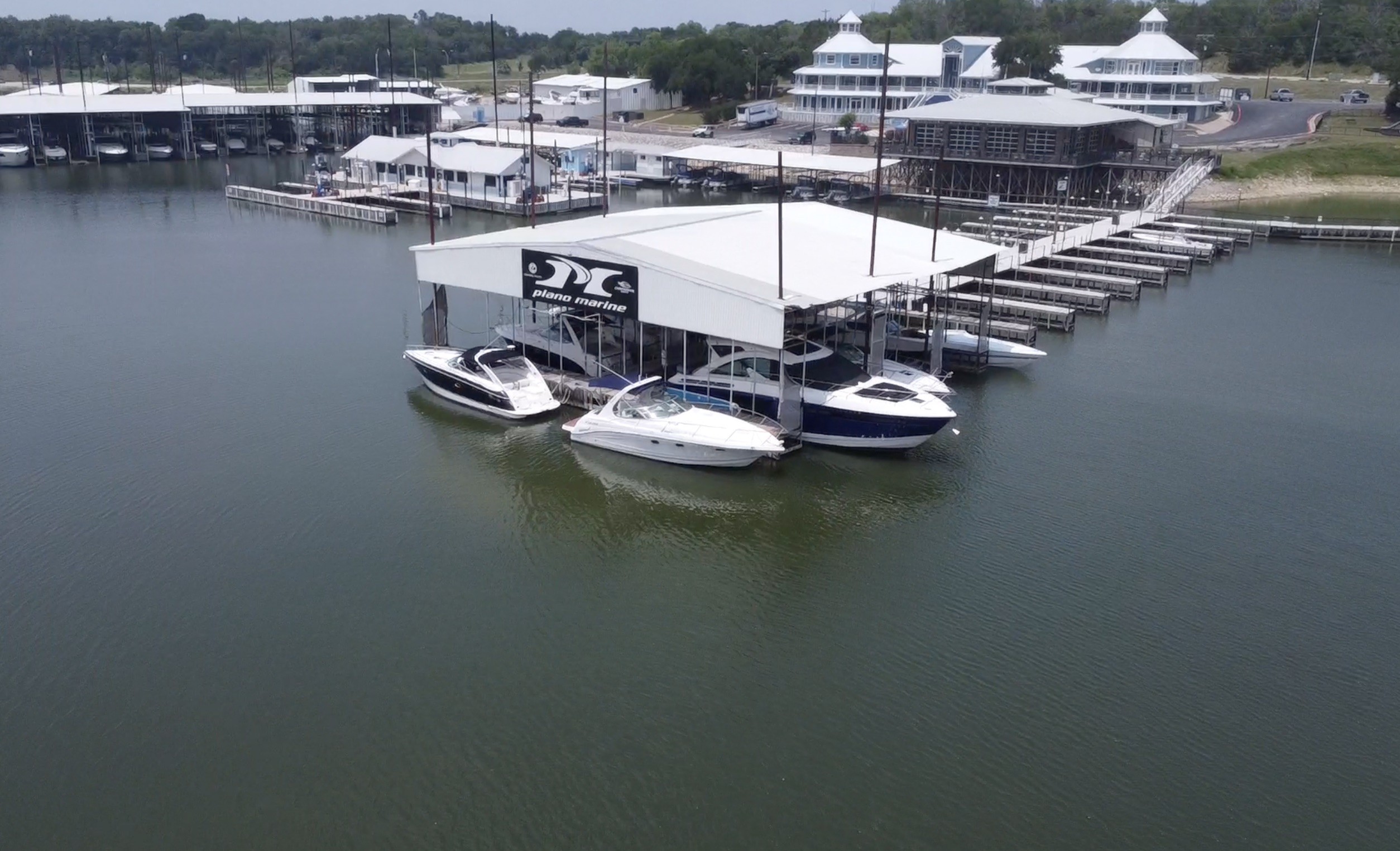 Aerial view of a marina with multiple white boats docked beside a large covered boat storage facility surrounded by calm waters and a backdrop of trees and buildings.