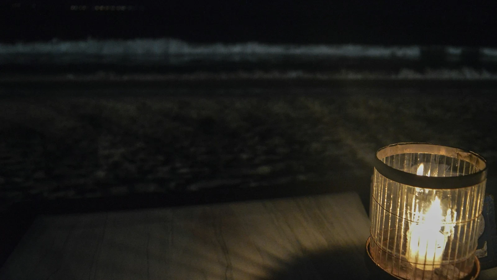 Ein Windlicht mit brennender Kerze steht am Abend auf einem Holztisch, im Hintergrund das Meer
