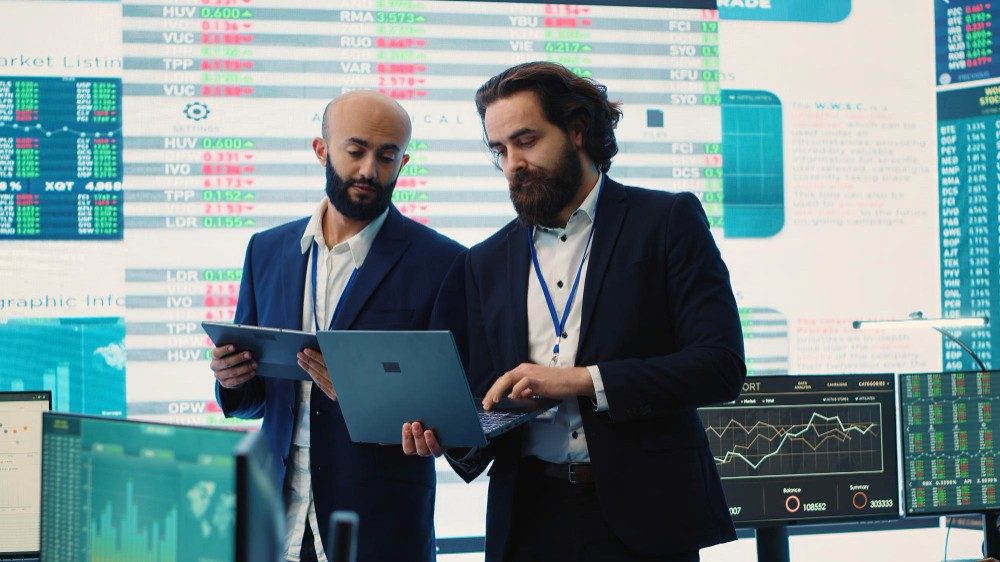 Two businessmen looking at a laptop. Behind them is a screen showing tables and charts of profit status.