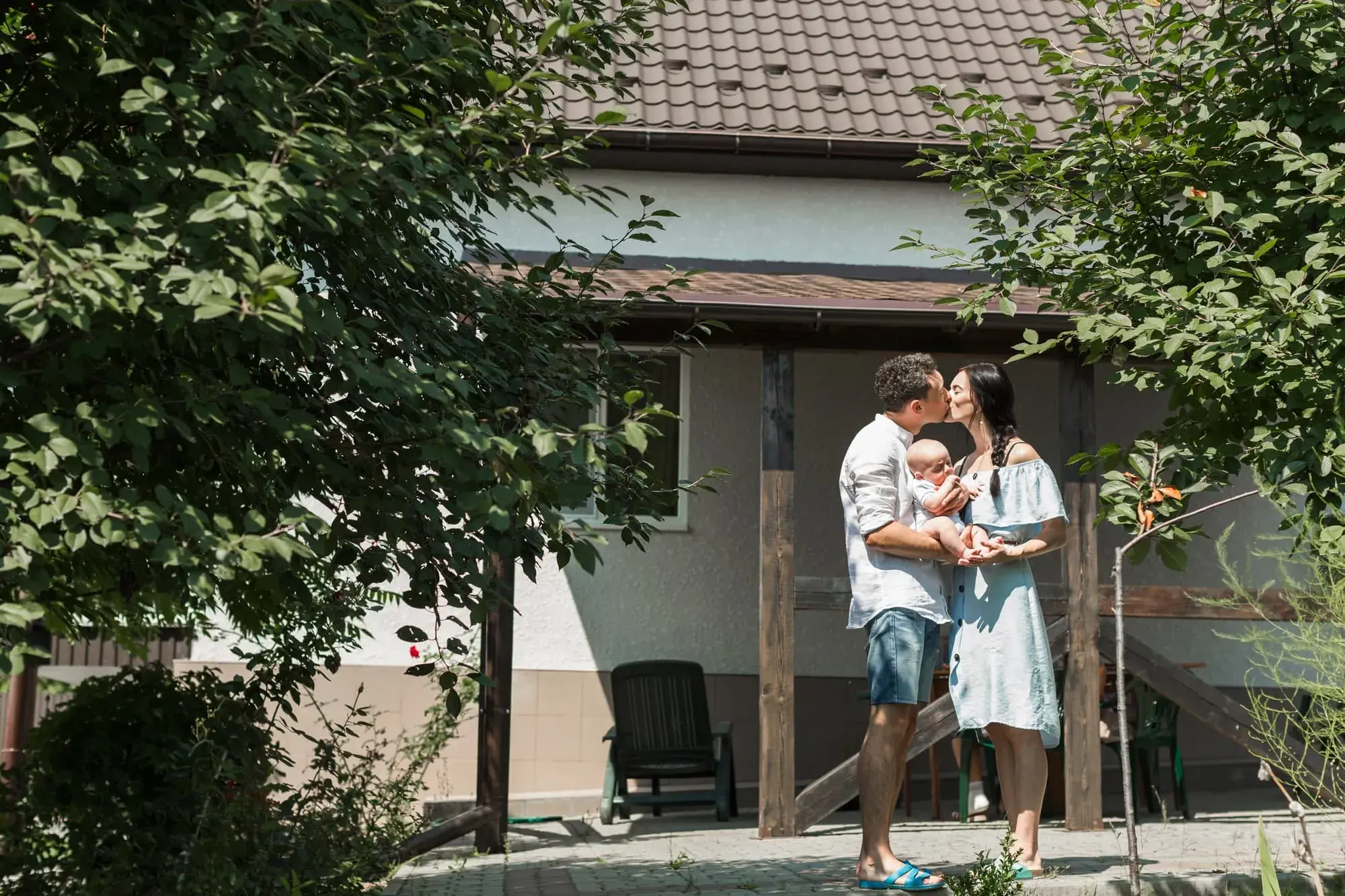 A young family stands in front of their new home, smiling and holding their baby together on a sunny day. The photo captures the joy and sense of accomplishment that comes with purchasing a first home through Chris Lewis Home Loans.