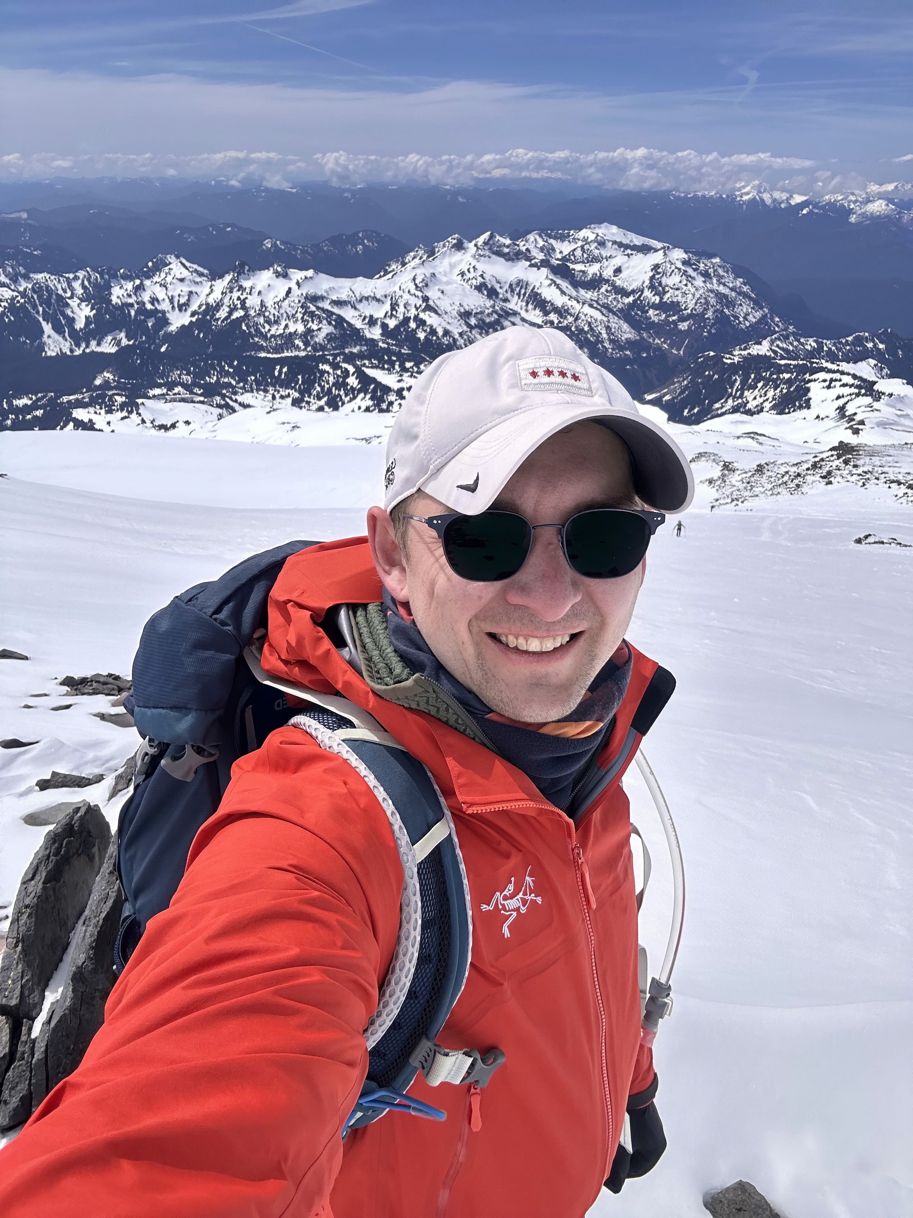 A smiling man with curly hair is sitting at a desk, looking at the camera, with a laptop and a lamp in the background.