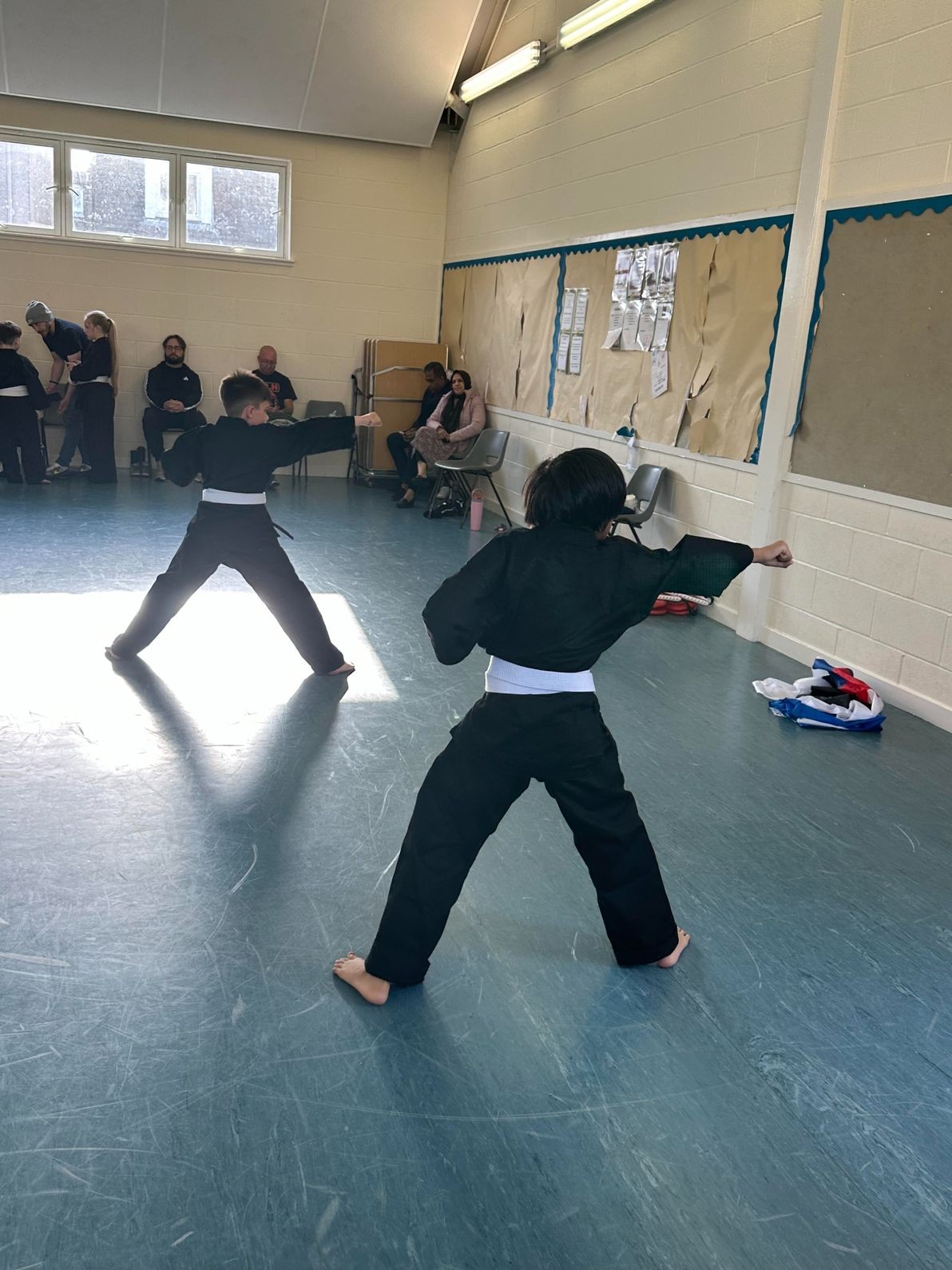 Two children in black martial arts uniforms and white belts practice patterns with focused expressions.