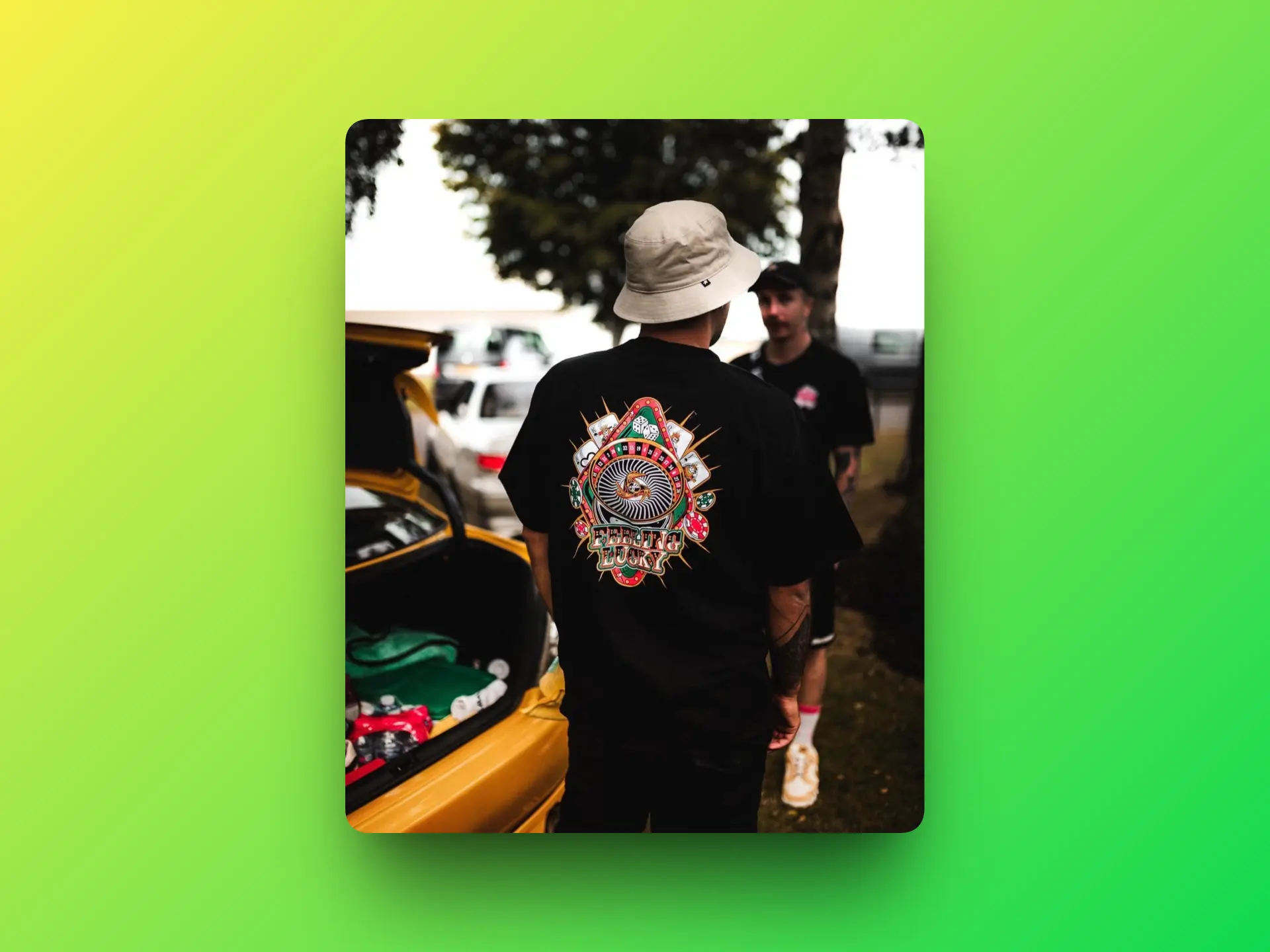 Person wearing a black Lowco Automotive “Feeling Lucky” graphic T-shirt with playing card and poker-inspired artwork, photographed beside a car at an outdoor automotive meet.