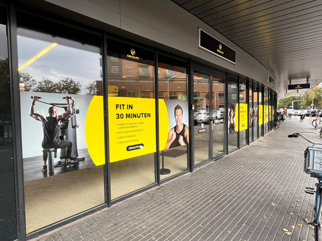 A Circle Fit hub with large glass windows featuring bold yellow signage and people visible indoors. Sidewalk in view.