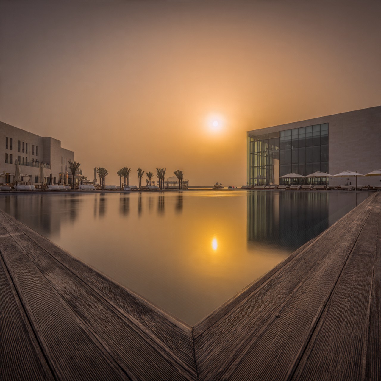 Sunset over a tranquil pool reflects warm orange hues. Modern buildings and palm trees frame the scene, exuding calm and peacefulness.
