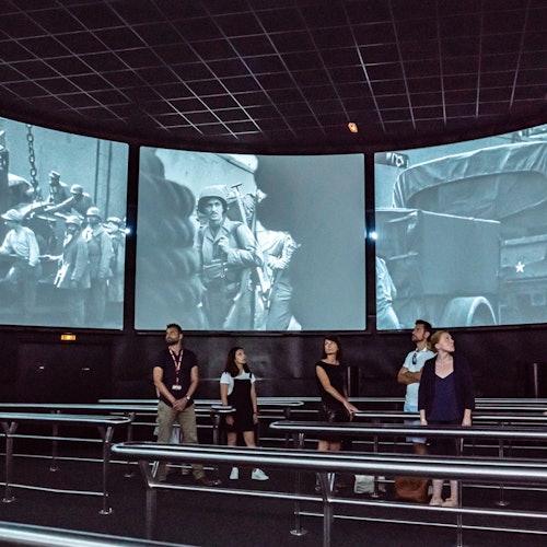 Menschen in einem dunklen Raum mit großen, wandmontierten Bildschirmen, die Schwarz-Weiß-Bilder aus Kriegszeiten zeigen.