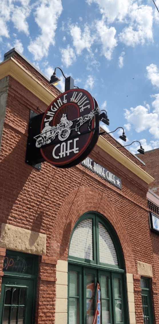 Exterior view of the Engine House Cafe with a large circular sign featuring an old fire engine, mounted on a brick building under a blue sky with clouds.